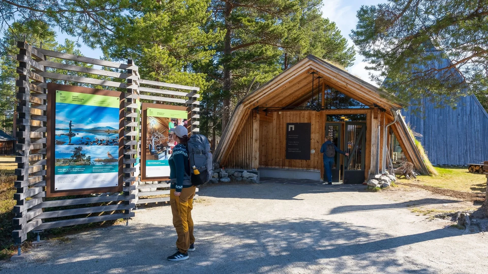 People visiting a nature park, looking at informational posters outside a wooden building surrounded by trees.