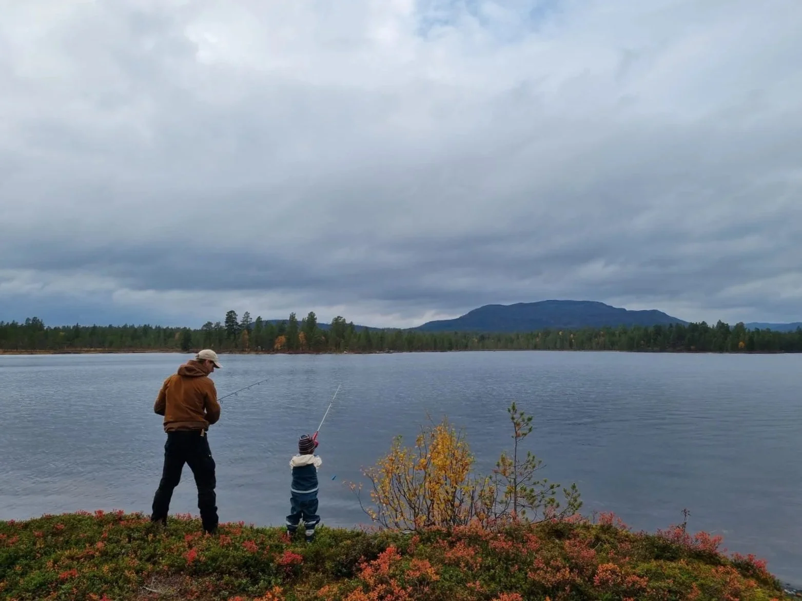A man and a child fishing by a lake with mountains and cloudy sky in the background.