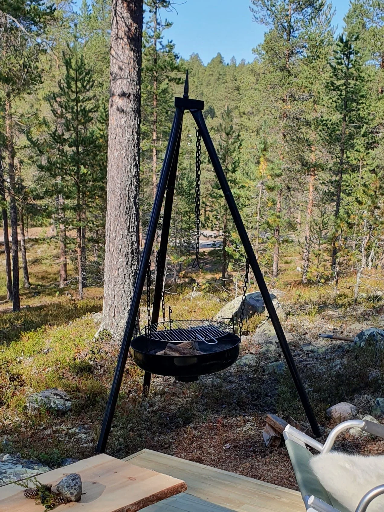 Ein schwarzer Dreibein-Lagerfeuergrill mit einem hängenden Rost und einer Pfanne, der auf einer Waldlichtung mit Bäumen im Hintergrund steht.