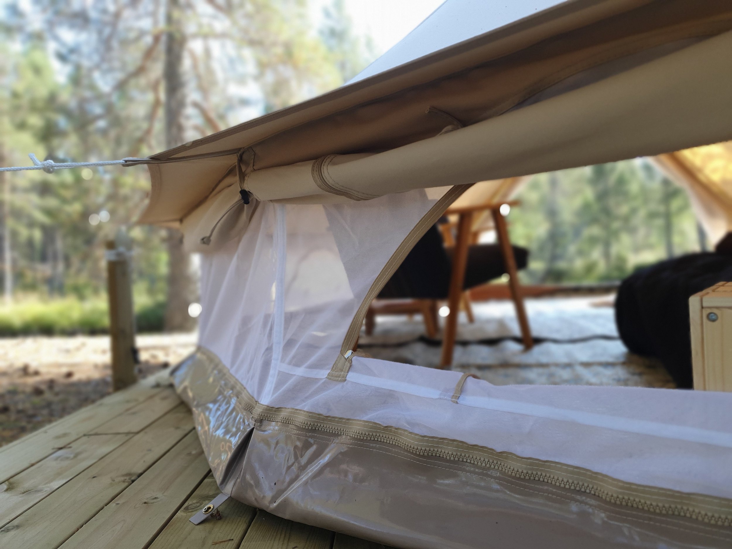 Glampingtelt med nettingklaff og glidelås på treterrasse, skogbakgrunn.