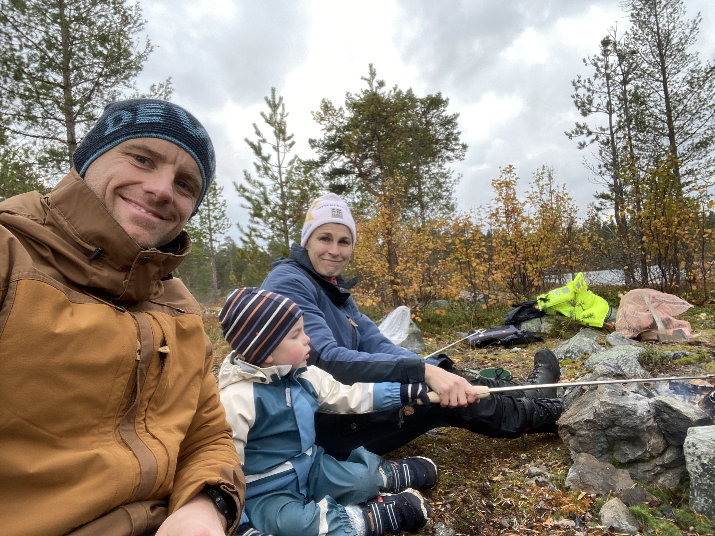 Family camping in a forest, man in brown jacket, child in blue snowsuit, woman cooking on campfire, trees in background.