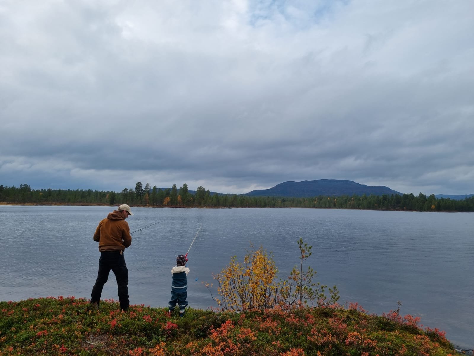 Two people fishing by a lake with a forest and mountain in the background, under a cloudy sky.