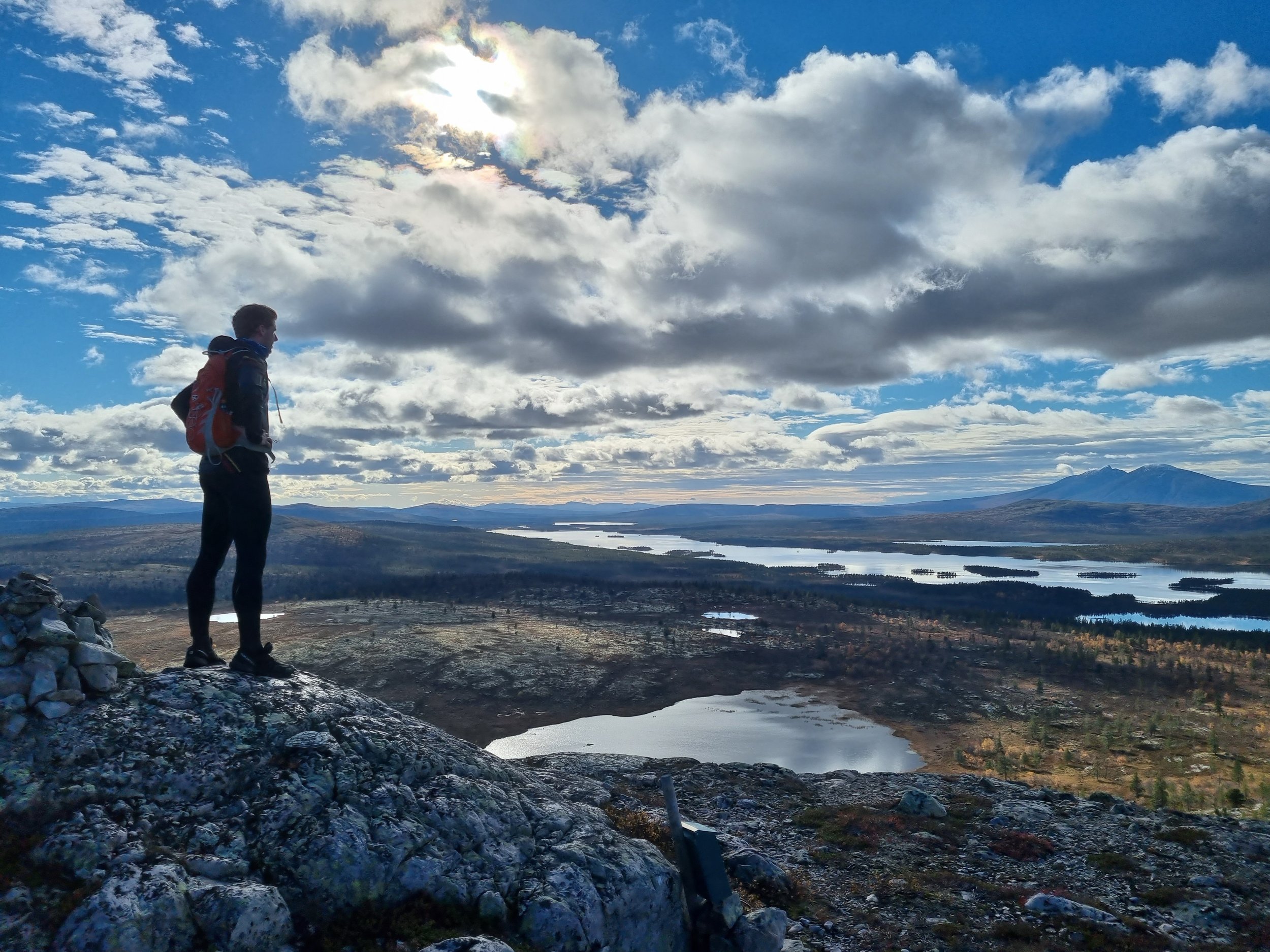 En turgåer med ryggsekk står på en steinete åstopp, med utsikt over et vidstrakt landskap med innsjøer, skoger og fjerne fjell under en delvis overskyet himmel.