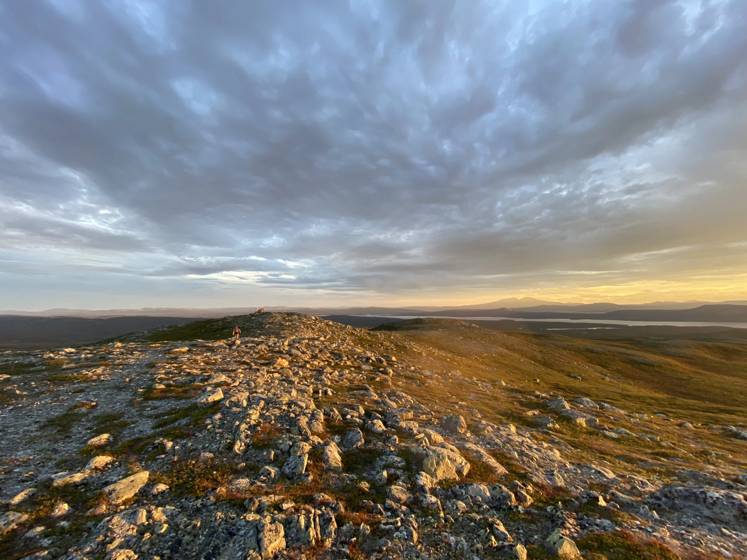Sunset over a rocky mountain landscape with a hiker in the distance and clouds in the sky.