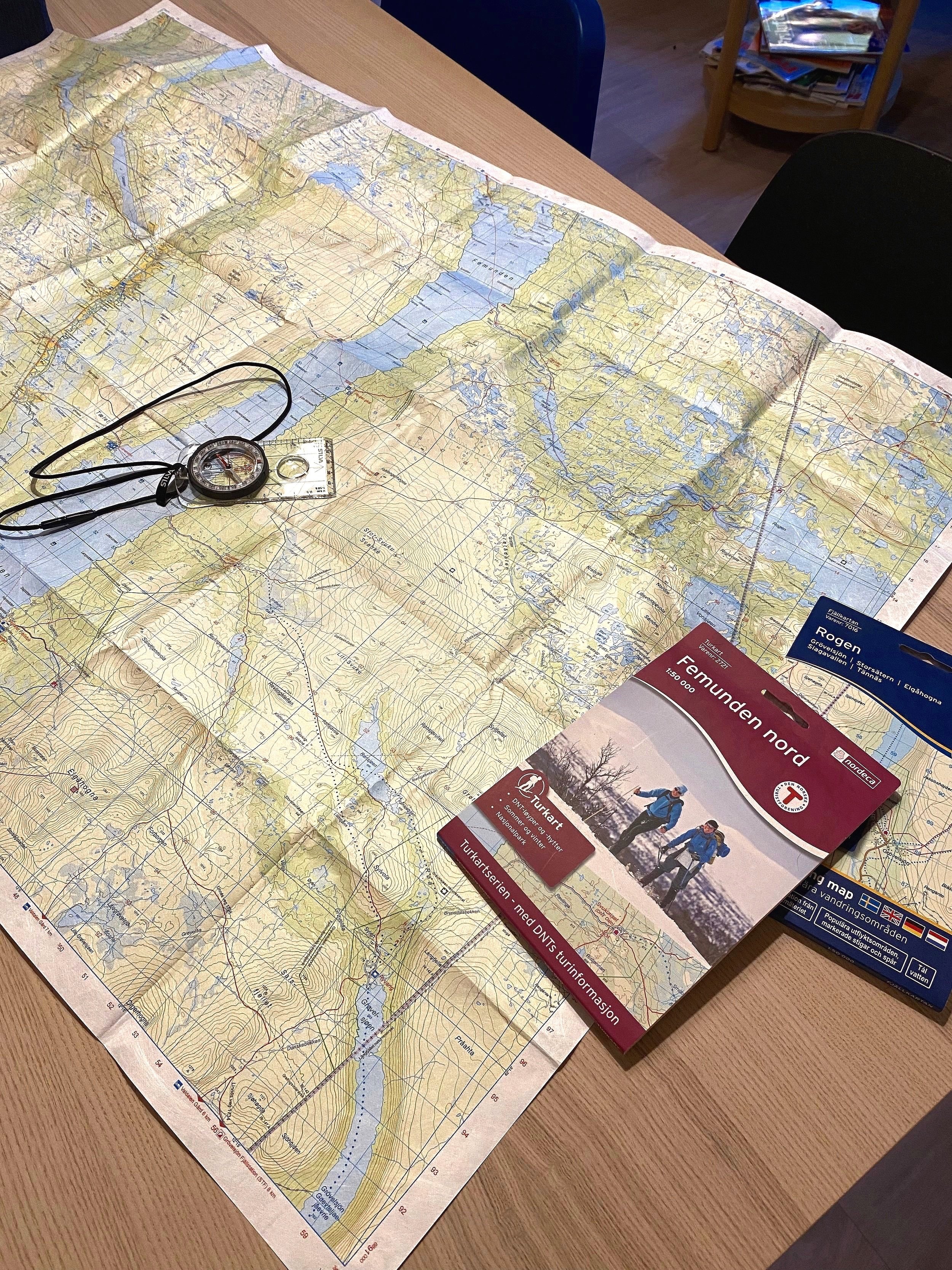 A topographic map spread out on a table with a compass on top, alongside a hiking guidebook labeled "Femunden nord" and a detailed walking map. The setting suggests planning for a hiking trip.