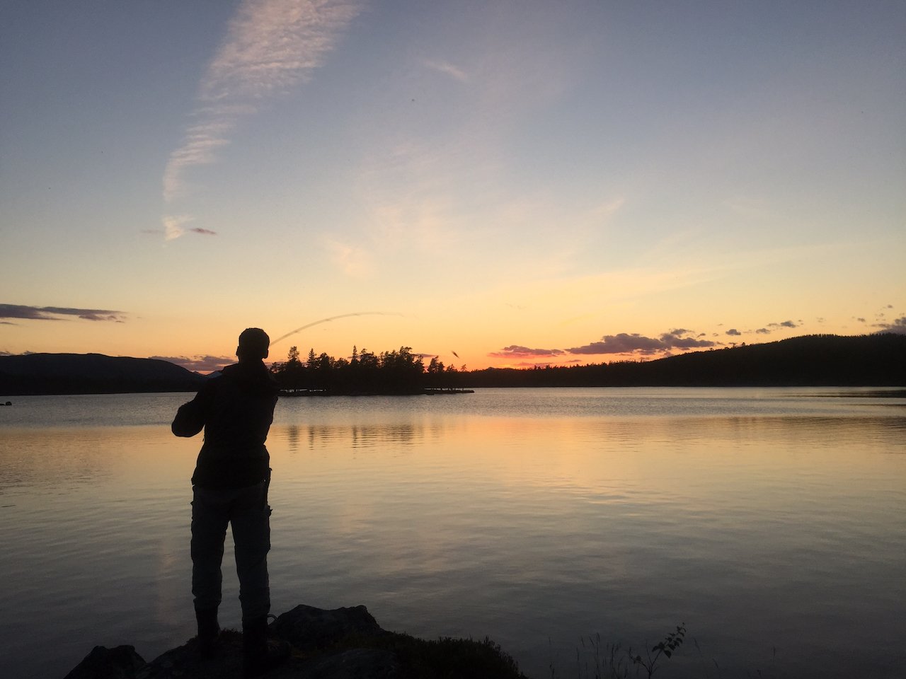 Silhuett av en person som fisker ved en rolig innsjø ved solnedgang med fargerik himmel og åser i bakgrunnen.