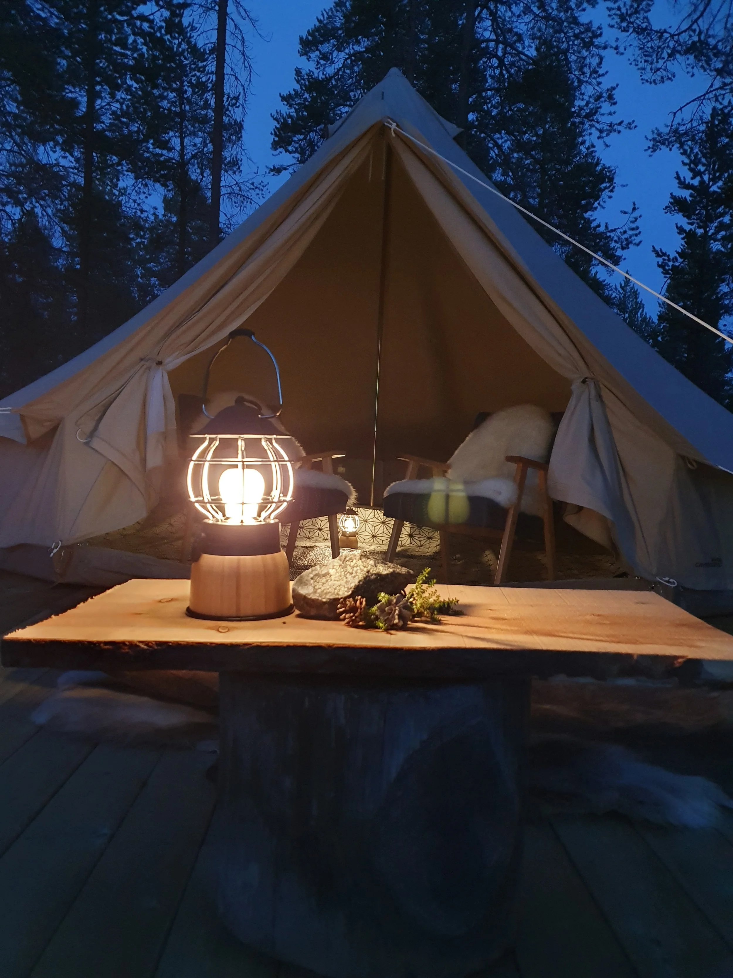 Eine leuchtende Laterne auf einem Holztisch vor einem Zelt in einem Wald in der Abenddämmerung.
