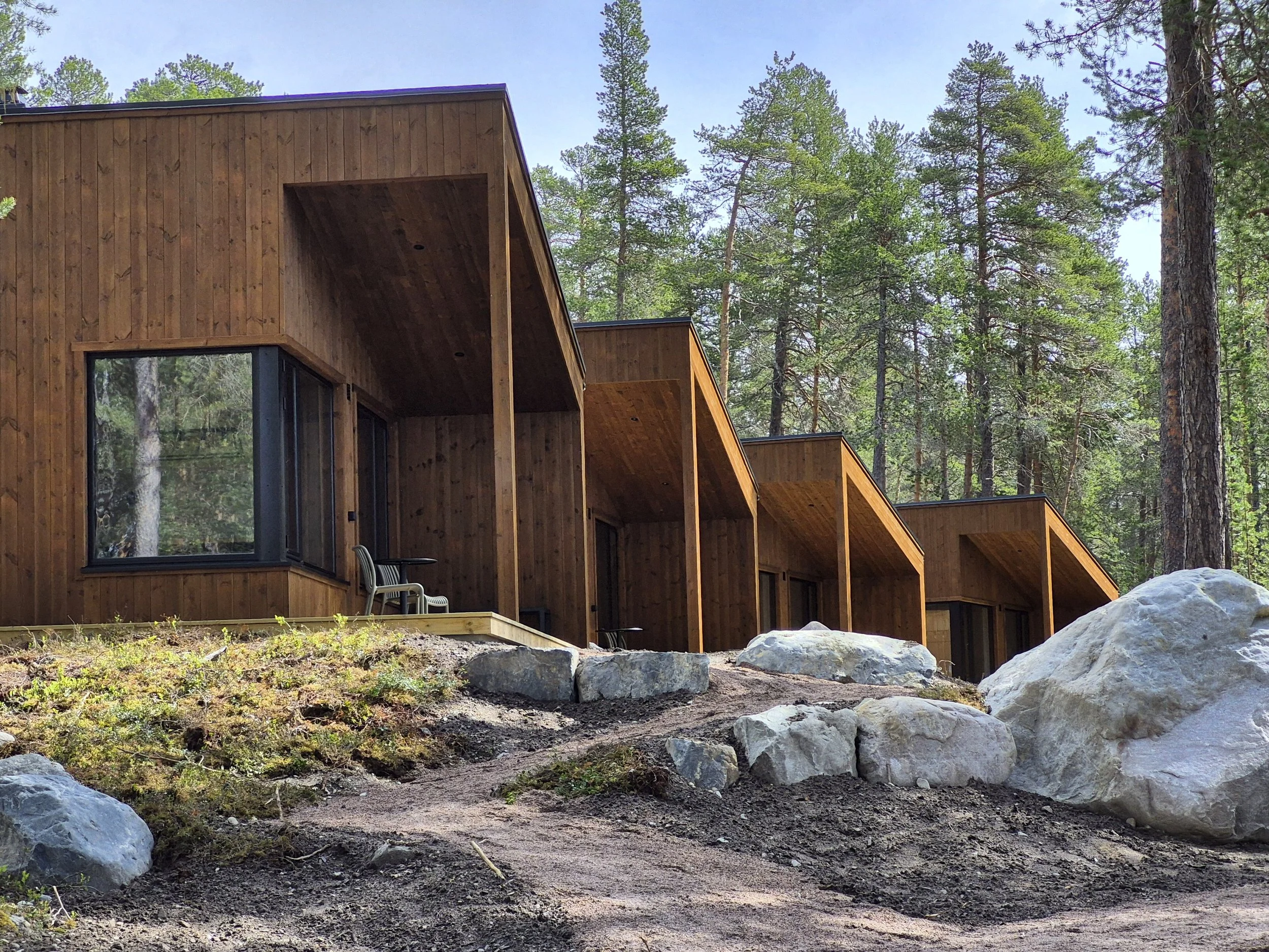 Three modern tiny houses with angular roofs set in a grassy and wooded area during sunset, with a campfire surrounded by logs in the foreground.