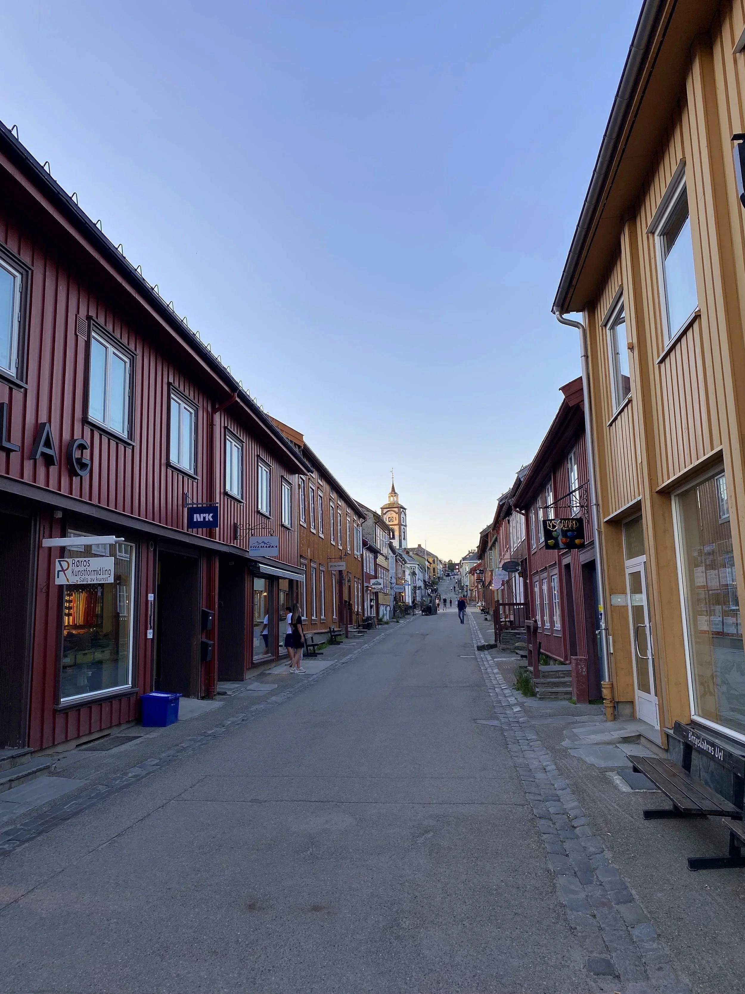 Empty street lined with colorful wooden buildings on both sides, with people walking, and a church steeple in the distance under a clear blue sky.