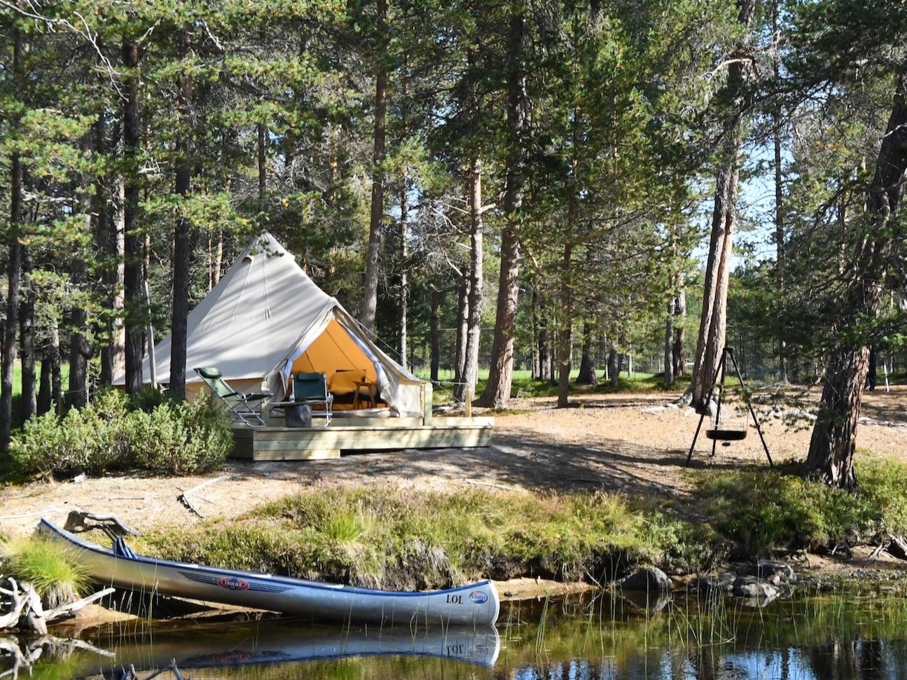 Camping scene with a large white tent on wooden platform in a forest, surrounded by tall trees. A canoe is on the shore of a lake, and there's a swing nearby.