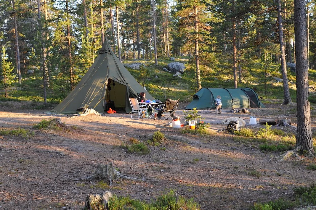 Camping scene in a forest with a teepee tent, a dome tent, folding chairs, a table, and camping supplies.