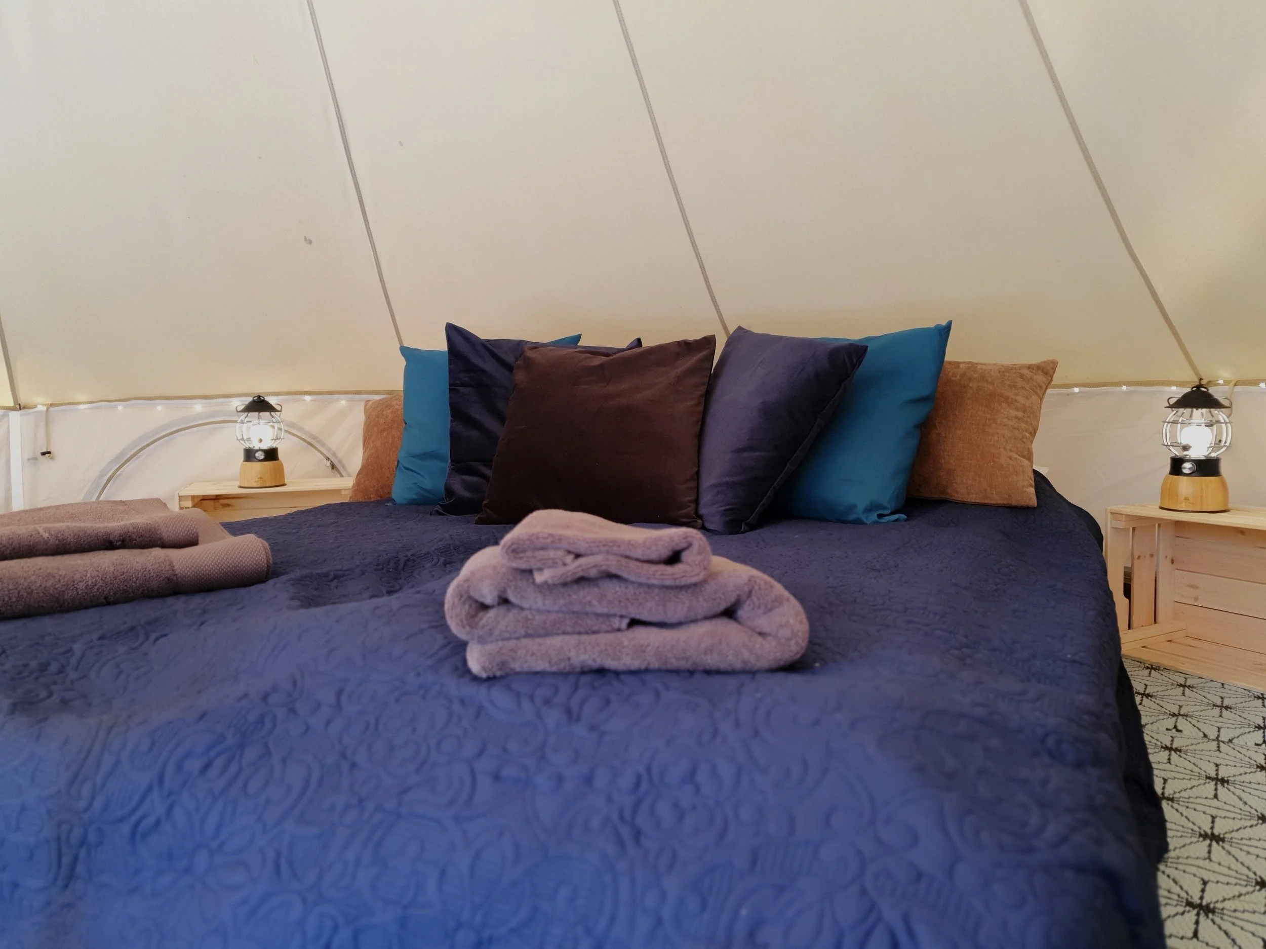 Innenansicht eines Zeltes mit einem ordentlich gemachten Bett, das mit einer blauen Steppdecke, mehreren bunten Kissen und gefalteten Handtüchern bedeckt ist. Zwei kleine Laternen stehen auf hölzernen Beistelltischen neben dem Bett. Das Zelt hat eine cremefarbene Innenausstattung und Lichterketten an der Seite.
