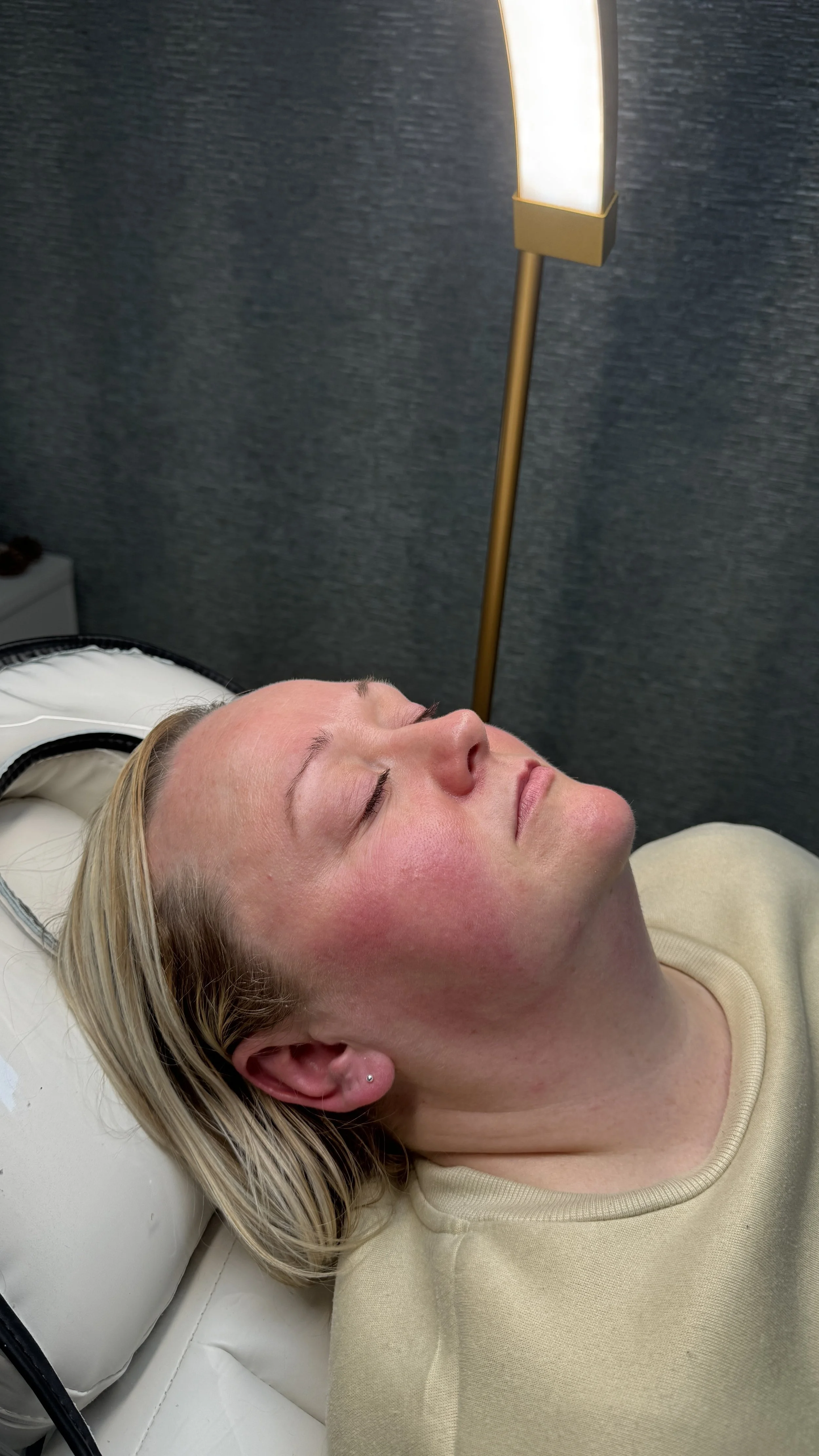 A woman relaxing with her eyes closed while lying on a bed or treatment table under a tall, modern floor lamp.
