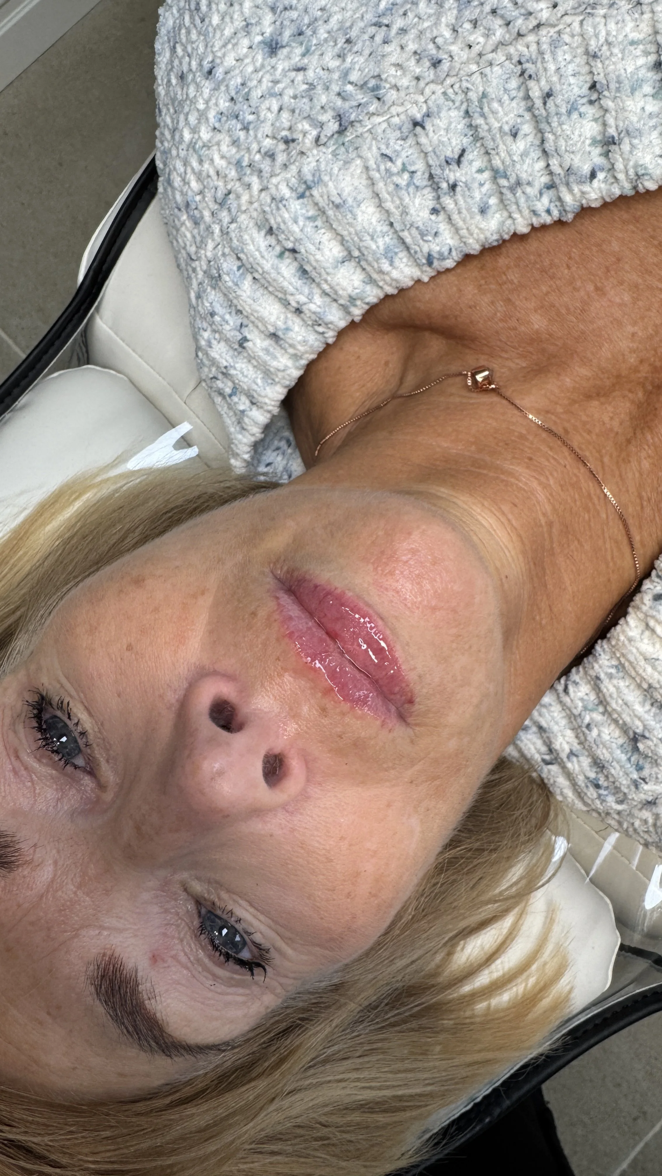 A woman with blonde hair lying down with her eyes open, showing her face, lips, and a necklace. She has a textured gray robe or towel draped over her shoulder.