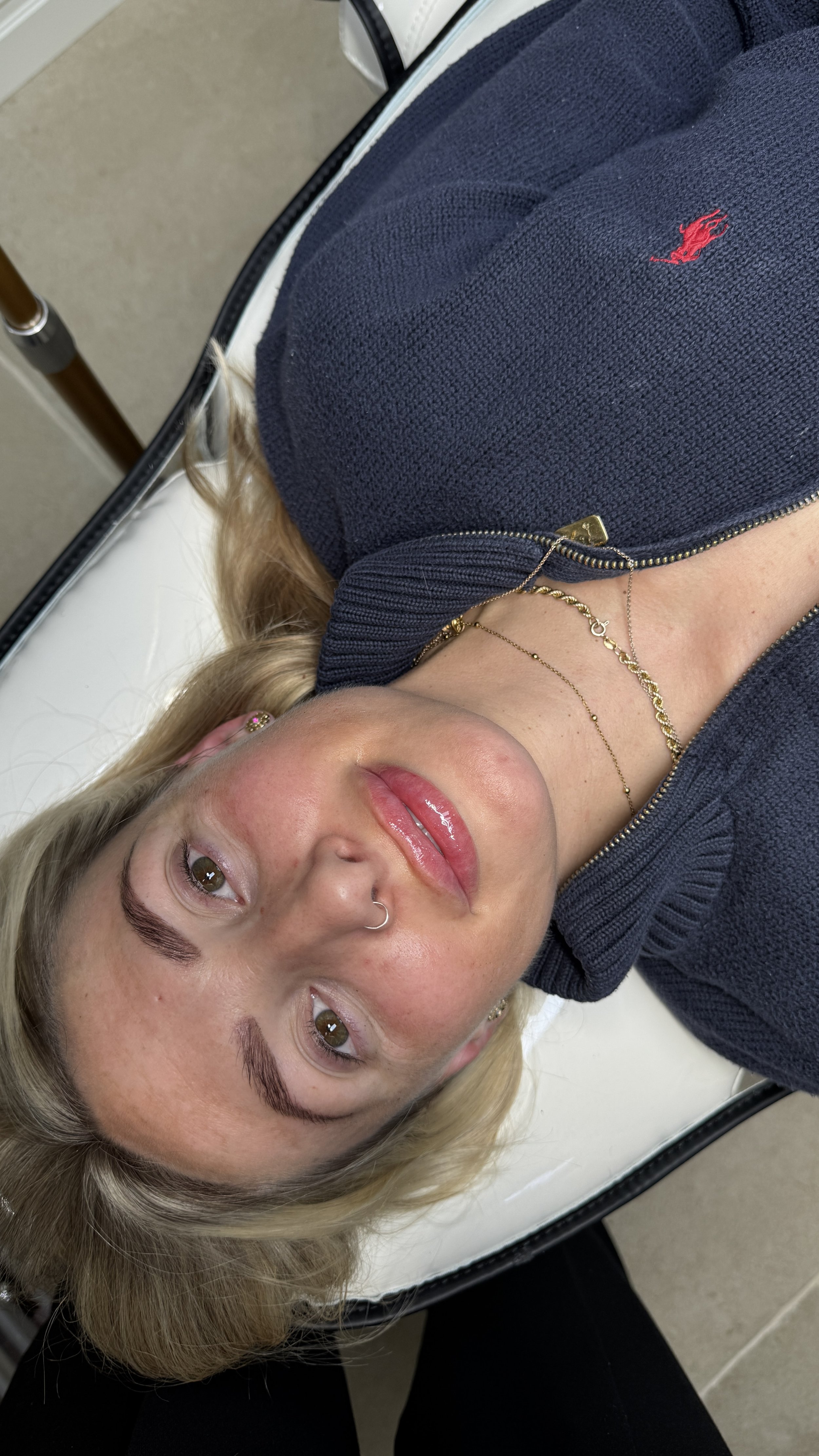 A woman with blonde hair, tan skin, and light makeup lying on a white cushioned surface. She is wearing a navy blue zip-up top with a red embroidered logo, layered gold necklaces, a nose ring, and gold earrings. The photo is taken from above with her