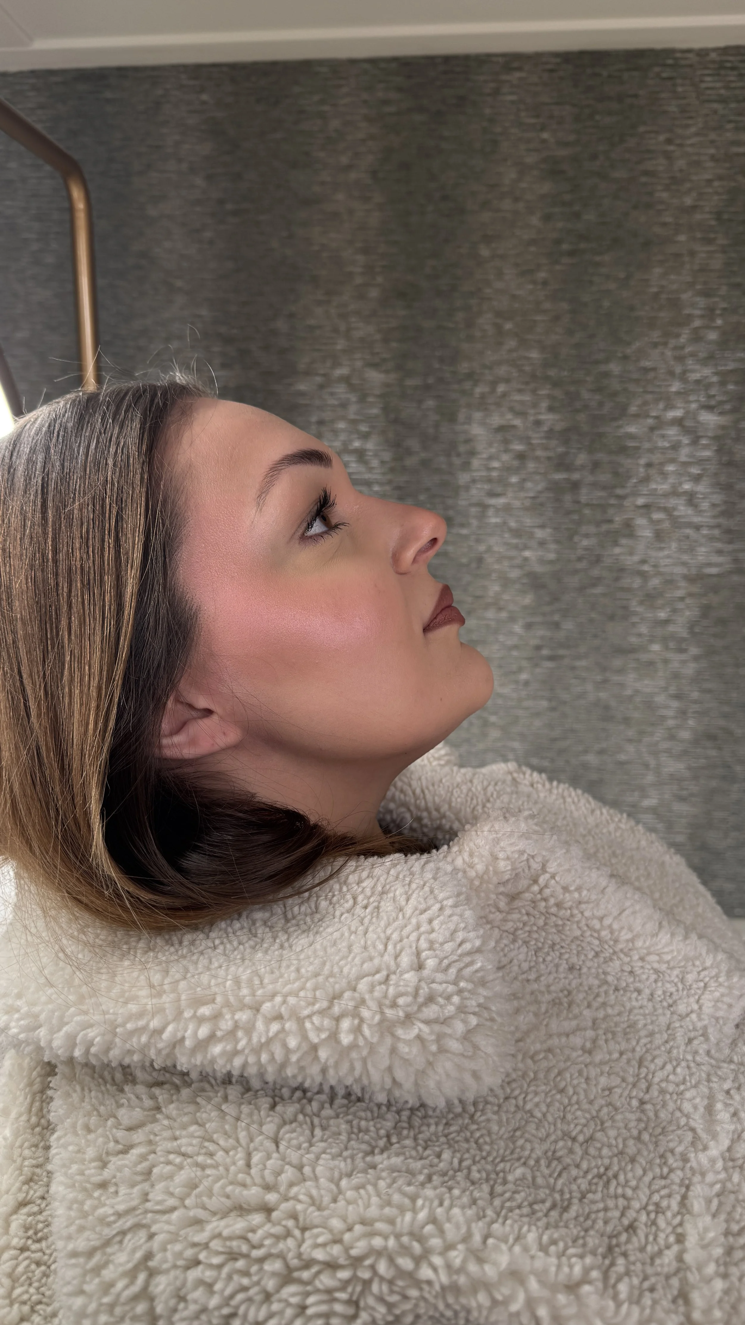 Side profile of a young woman with long brown hair, lying down with her head tilted back, covered in a fluffy beige blanket.
