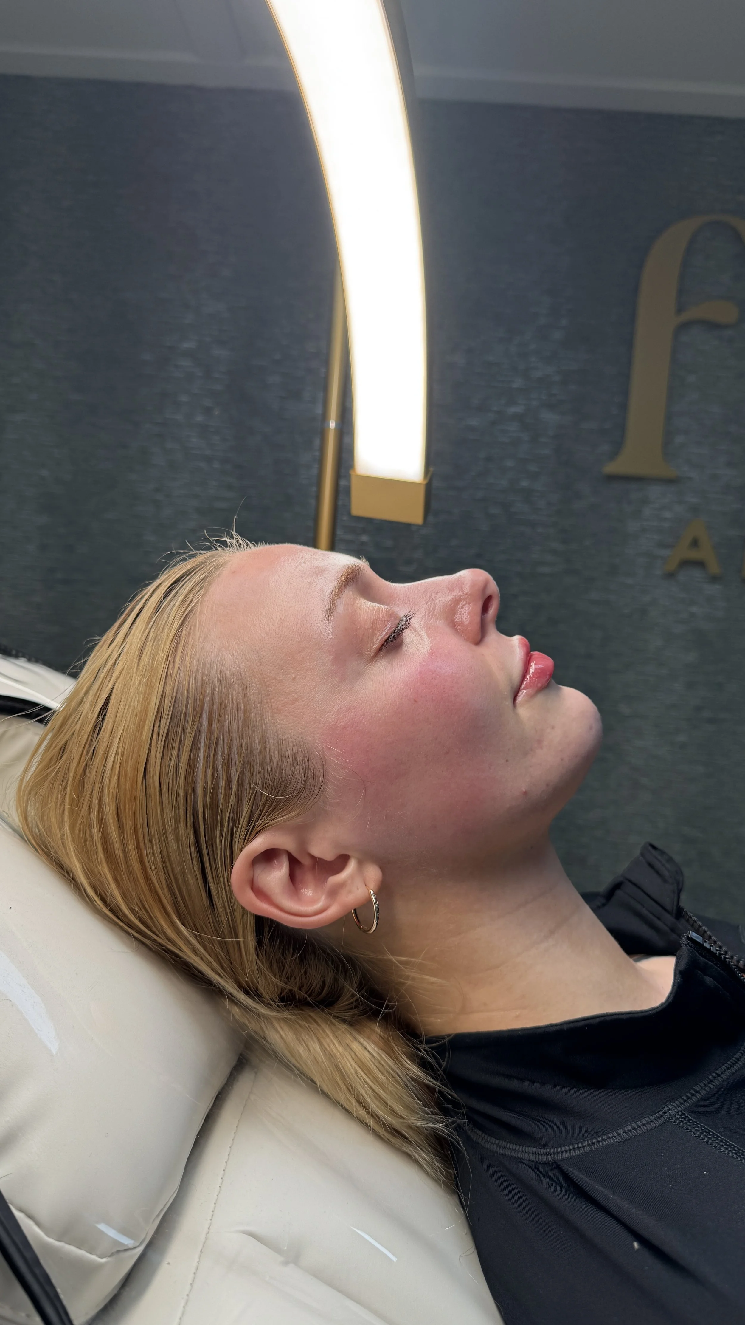 A young woman with blonde hair lying down with her eyes closed, head tilted back, and a relaxed expression, in a modern interior space with a blue textured wall and a large illuminated light fixture.
