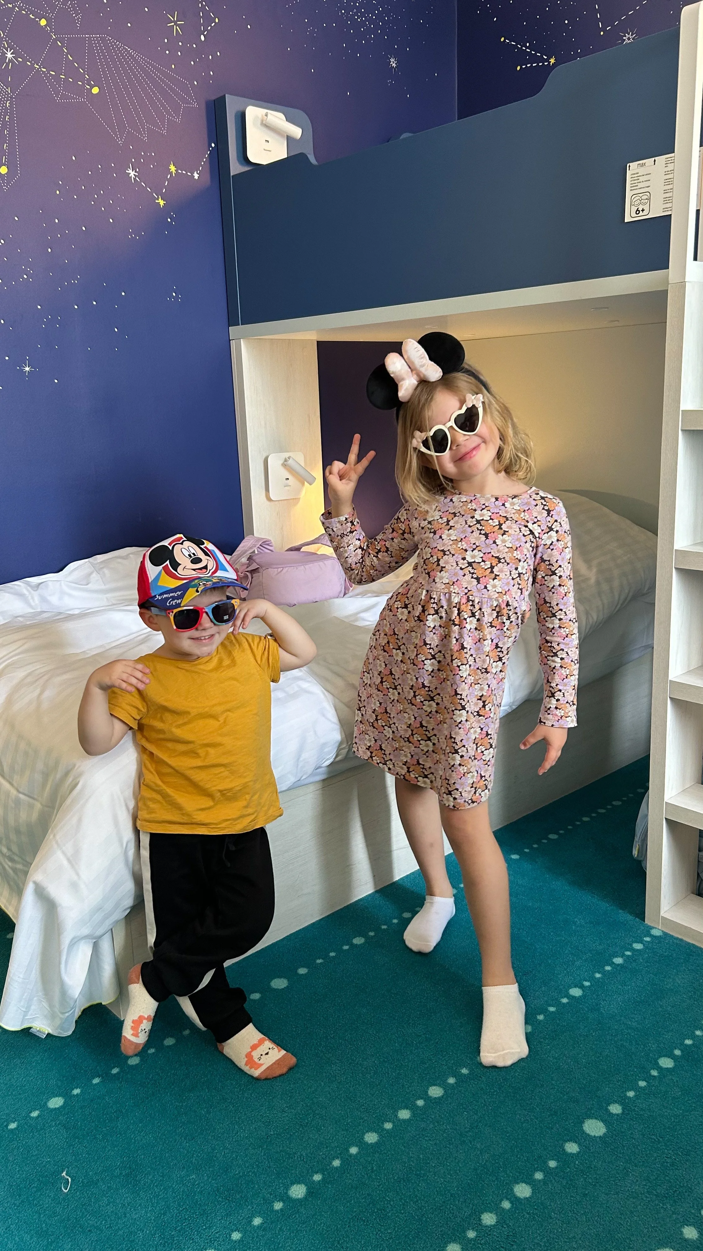 Two children in sunglasses and playful outfits posing on a hotel bed, with a starry sky mural on the wall behind them.