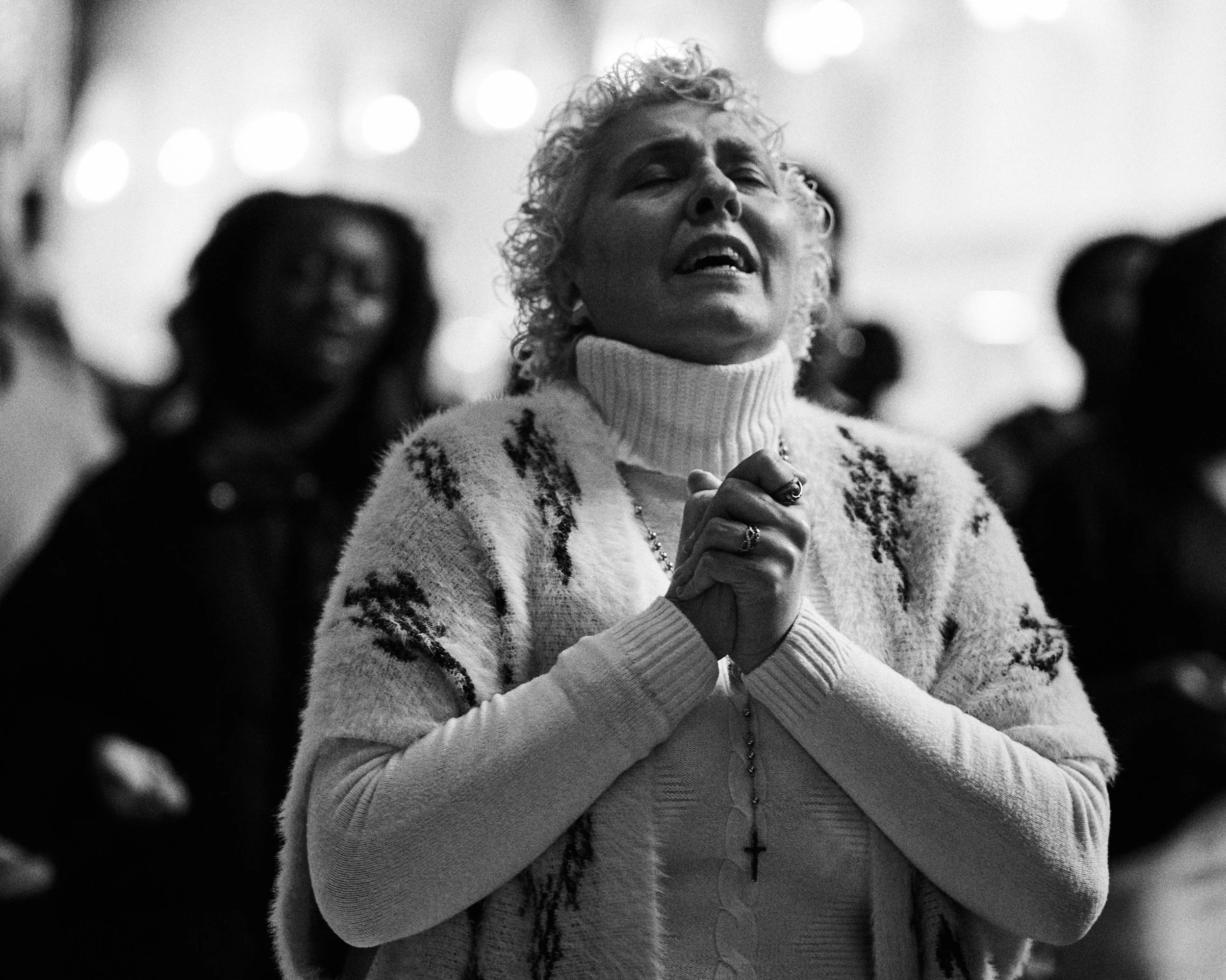 Une femme âgée aux cheveux courts et bouclés, vêtue d'une écharpe blanche et d'un pull à motifs, tient ses mains jointes en prière avec un rosary, dans un lieu semble-t-il de culte, entourée de plusieurs personnes dont une en arrière-plan.