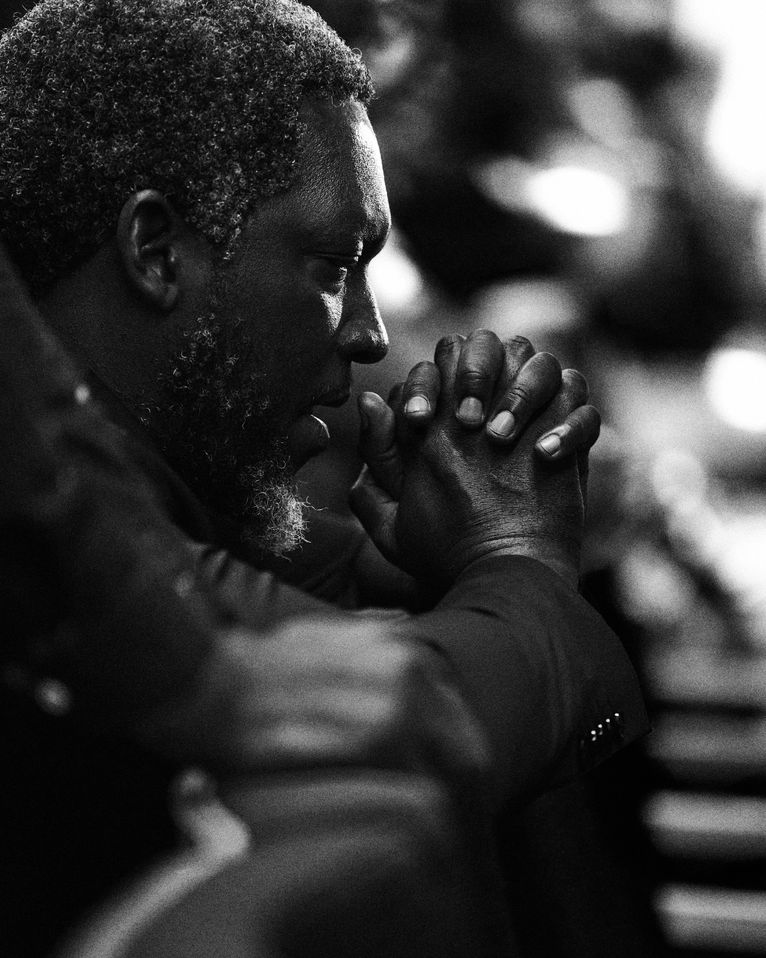 Un homme noir aux cheveux gris, les mains jointes en prière, les yeux fermés, dans une posture de méditation ou de recueillement, en noir et blanc.