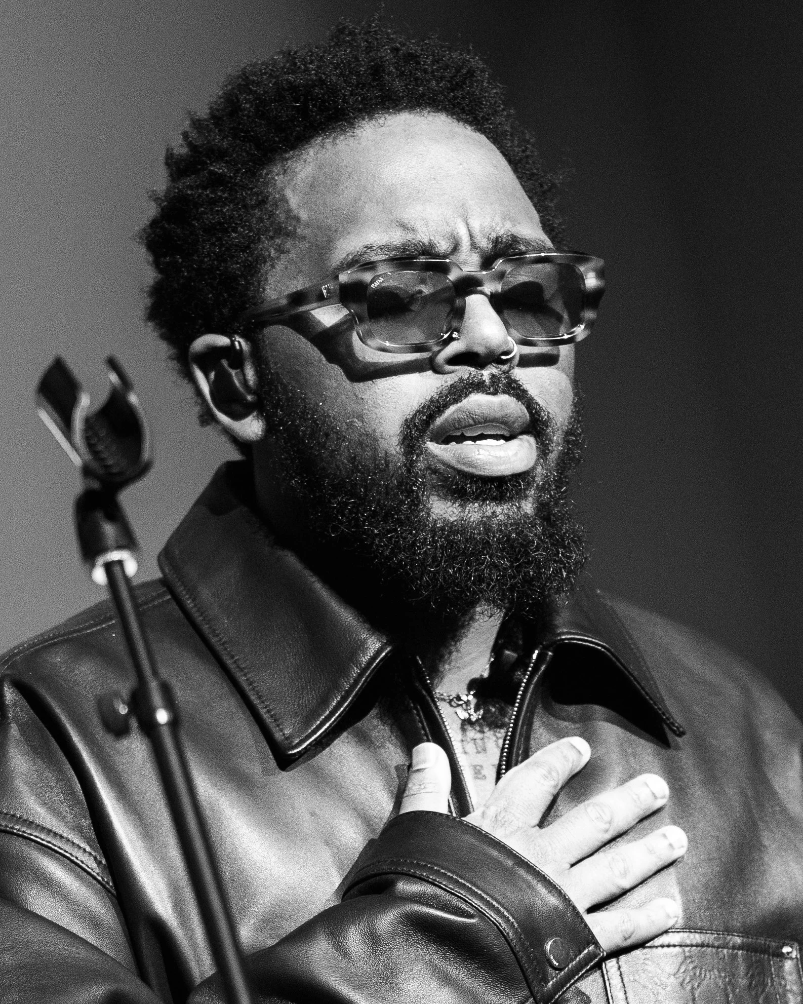 Un homme avec des lunettes épaisses, une barbe et une coiffure afro, portant une veste en cuir, tenant sa main sur sa poitrine dans une expression intense, en noir et blanc.