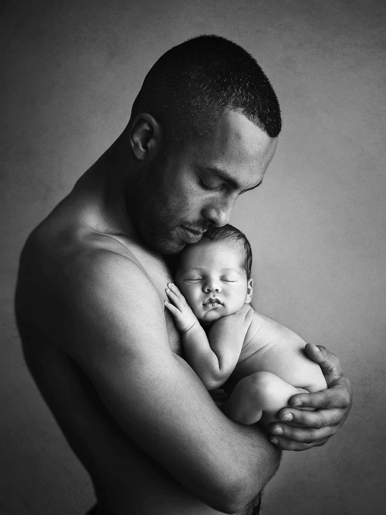 A black and white photograph of a shirtless man holding a sleeping baby close to his chest. The man is looking down gently at the baby, who is resting peacefully with eyes closed and face nestled against the man's chest.