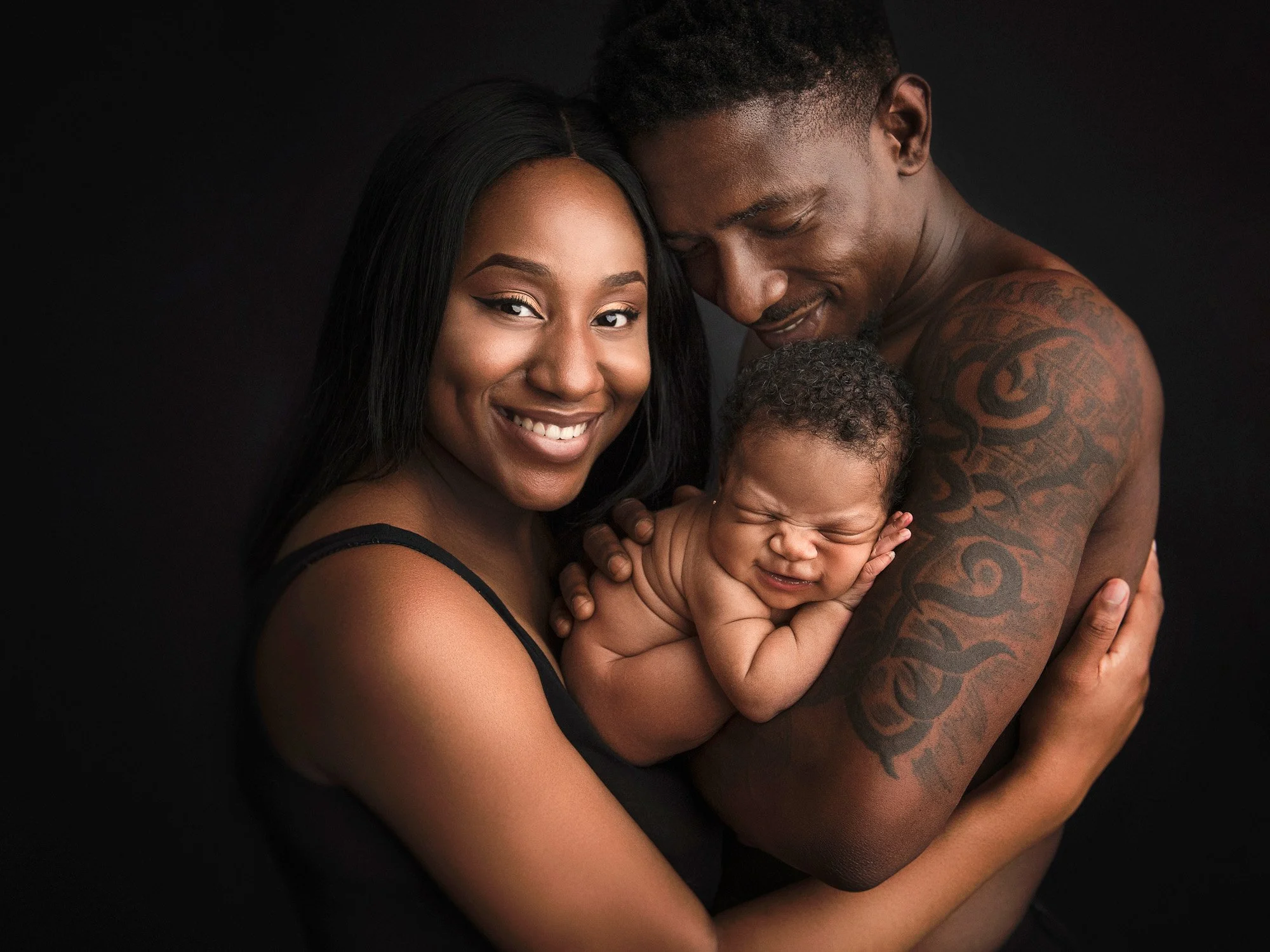 A happy Black family of three holding a newborn baby against a dark background. The mother is smiling, the father is resting his head on the baby's head, and the baby is squinting with a playful expression.