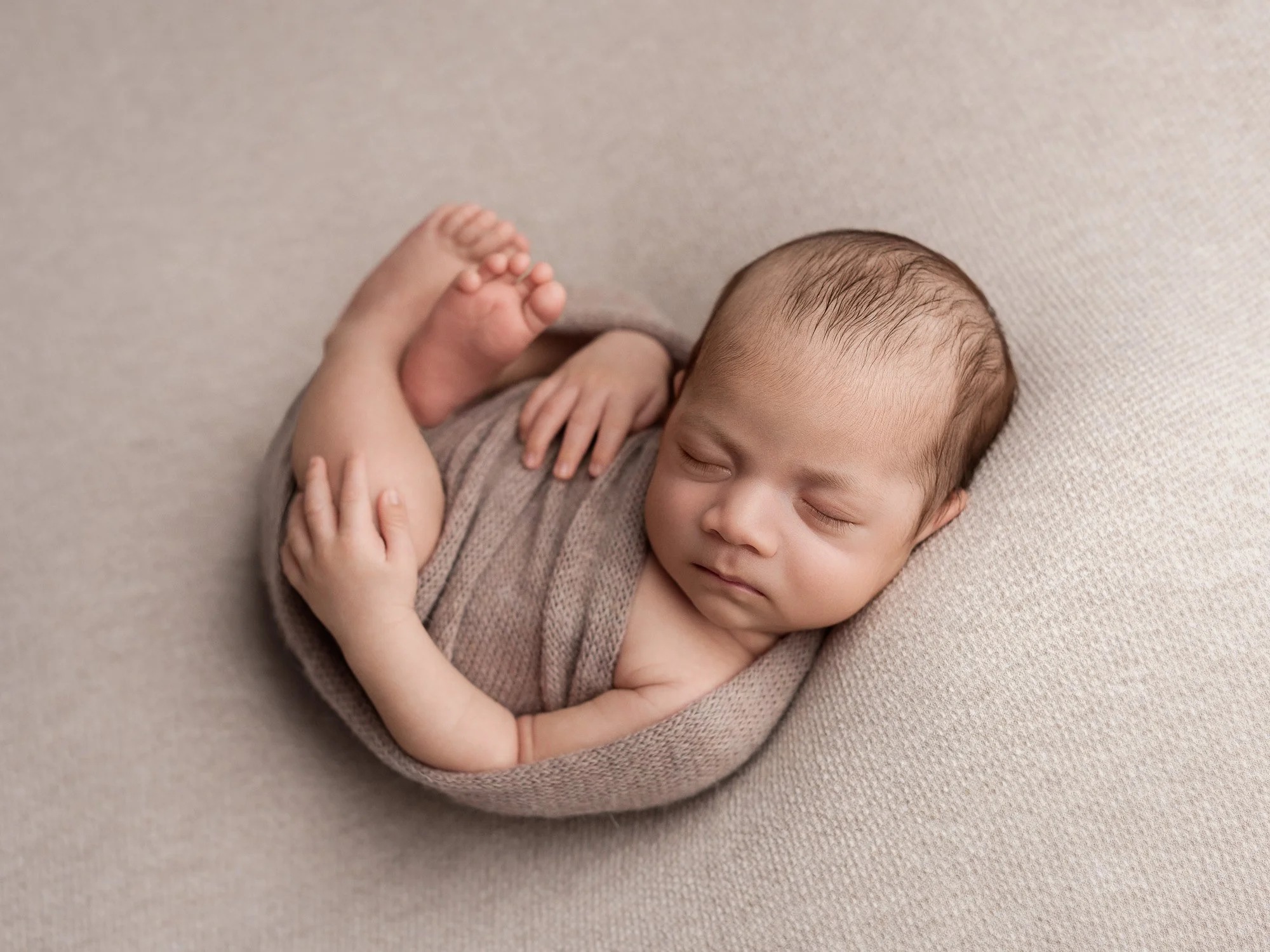 A sleeping newborn baby wrapped in a beige blanket, lying on a beige textured surface.