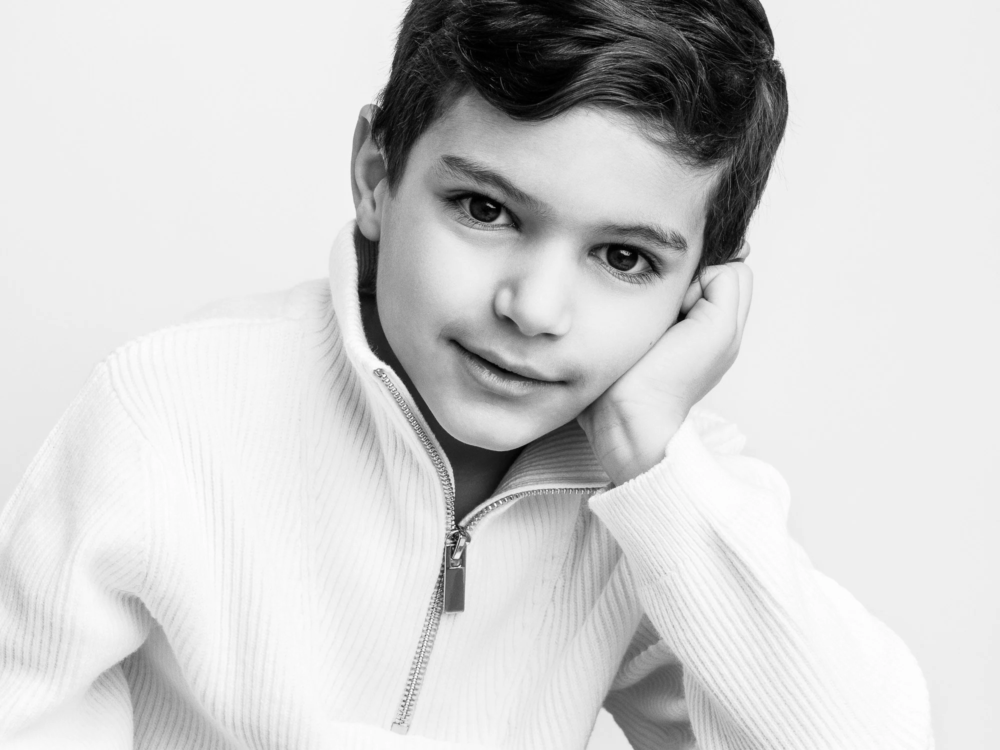 A black-and-white portrait of a young boy with short dark hair, wearing a zip-up sweater, resting his head on his hand, and smiling softly at the camera.