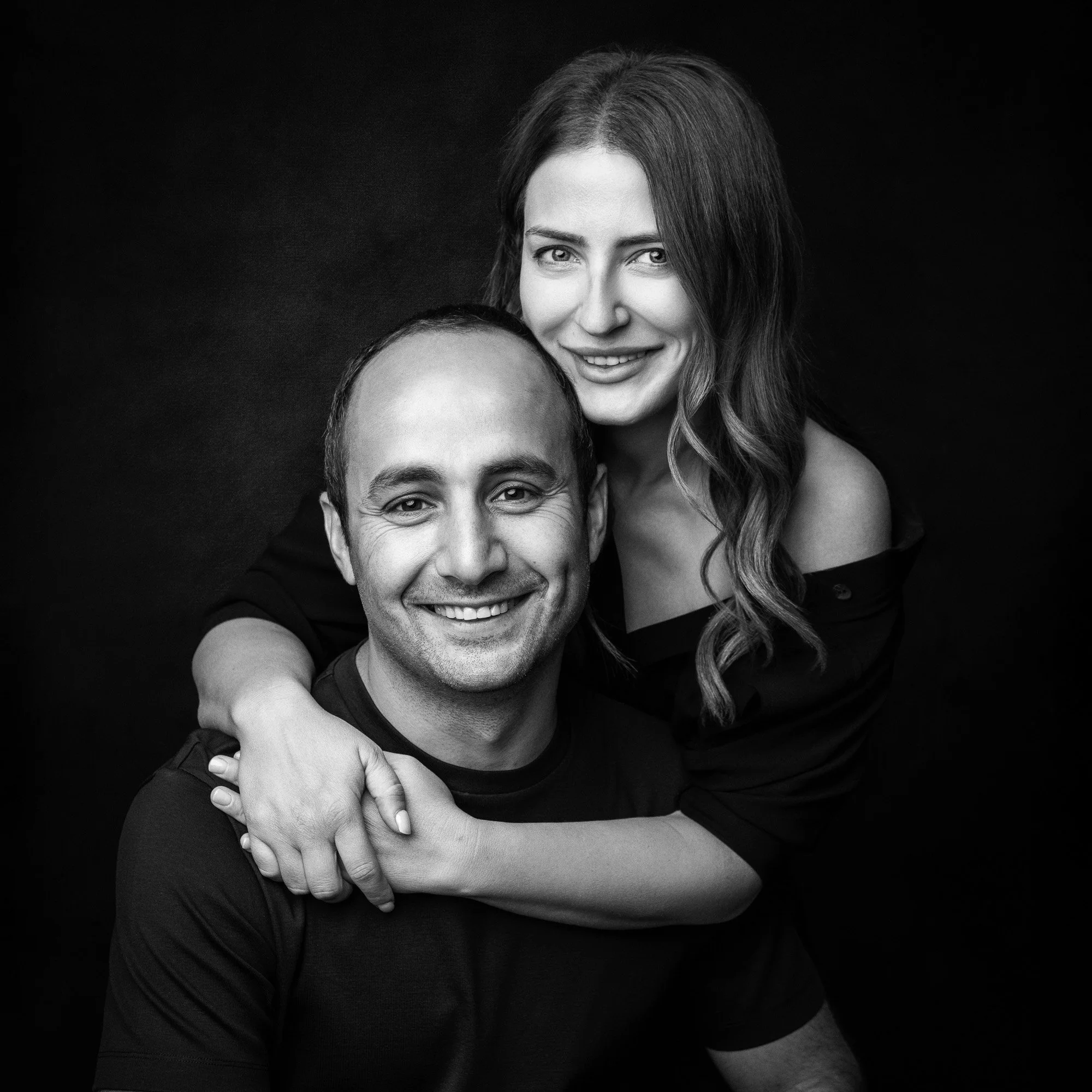 Black and white portrait of a smiling couple, the woman embracing the man from behind with her arm around his shoulder against a dark background.