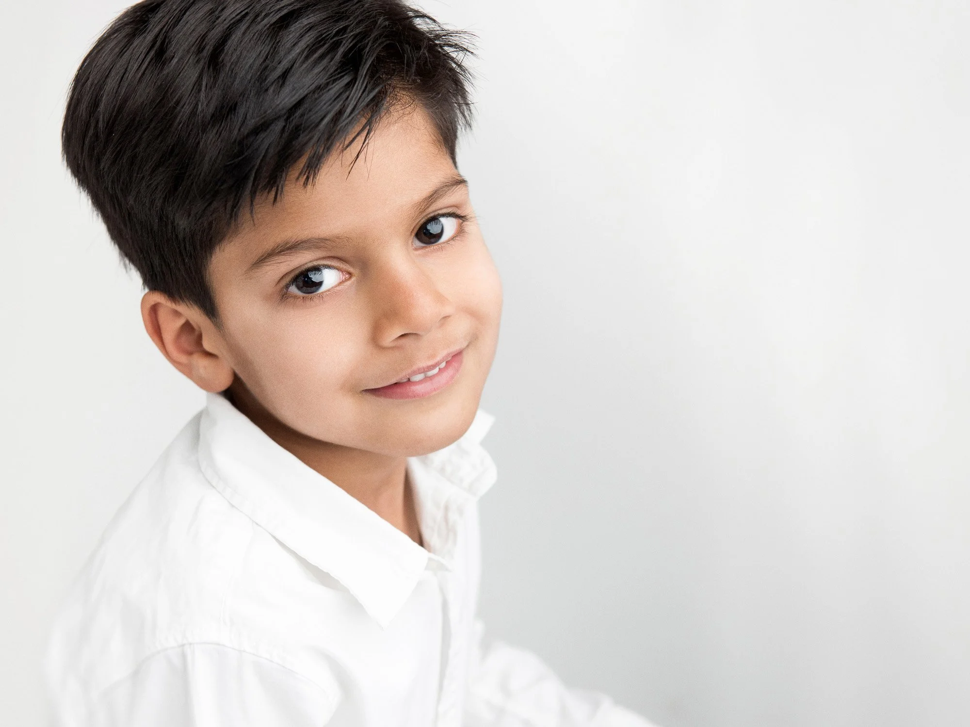 A young boy with dark hair and brown eyes wearing a white shirt, smiling gently, against a plain white background.