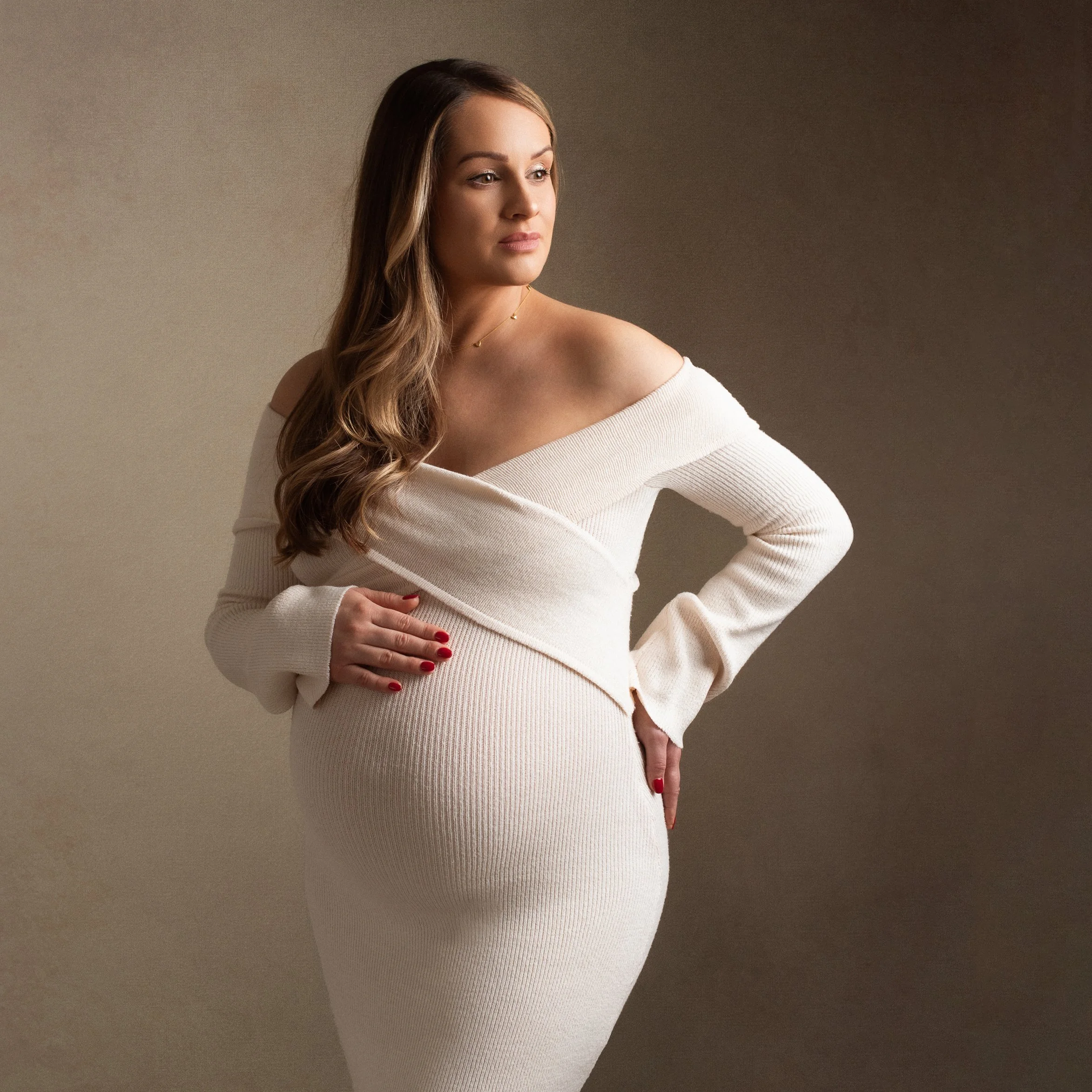A pregnant woman with long brown hair in a white off-shoulder dress posing against a beige background.