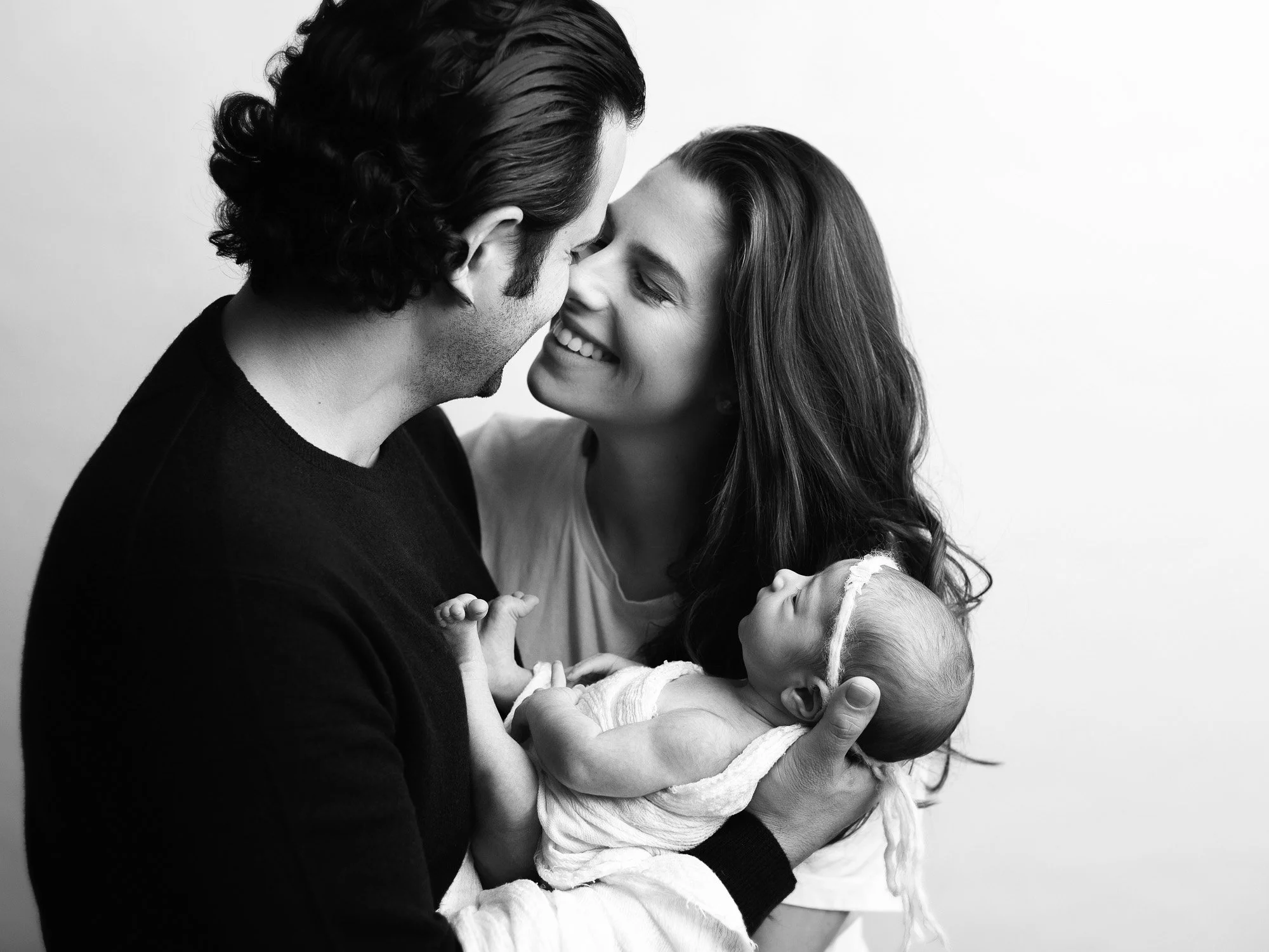 A black and white photo of a smiling woman holding a newborn baby, with a man close by, touching foreheads and sharing a joyful moment.