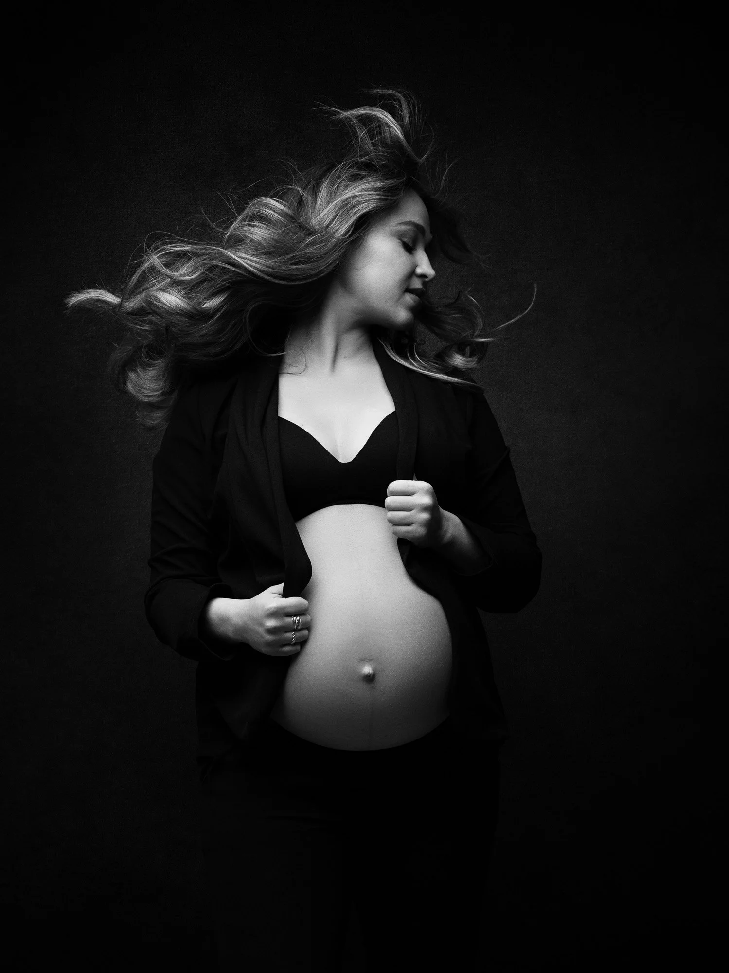 Black and white photo of a pregnant woman with long wavy hair, pose with her eyes closed, wearing a black bra and open black shirt, holding the shirt around her belly against a dark background.