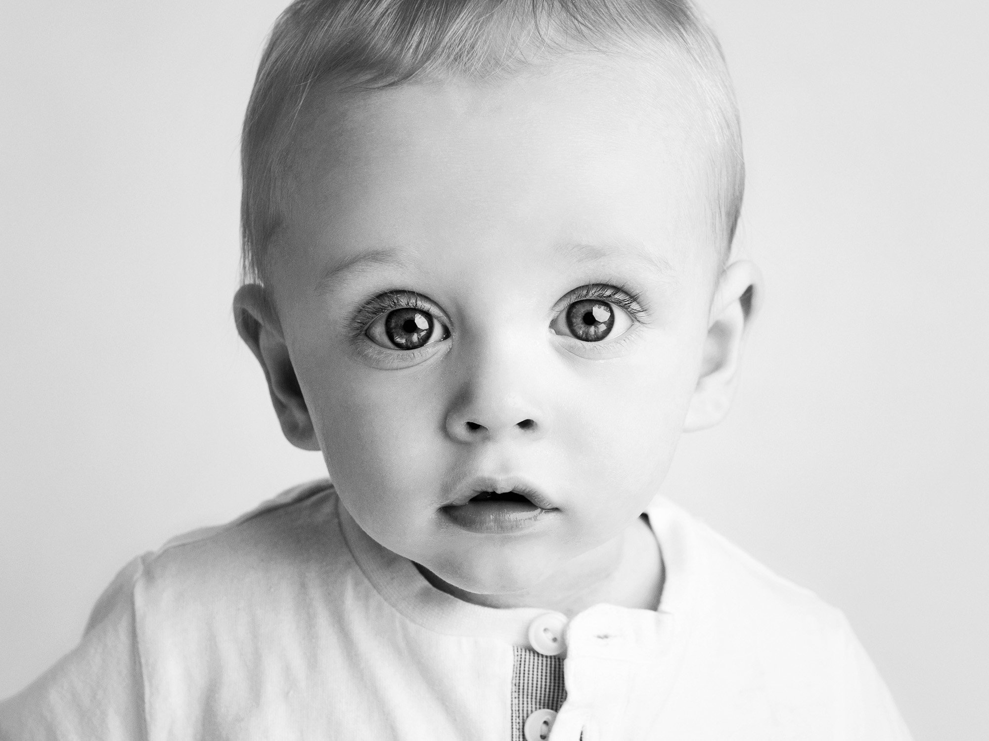 Black and white portrait of a young child with wide eyes, slightly open mouth, wearing a light-colored shirt, looking directly at the camera.