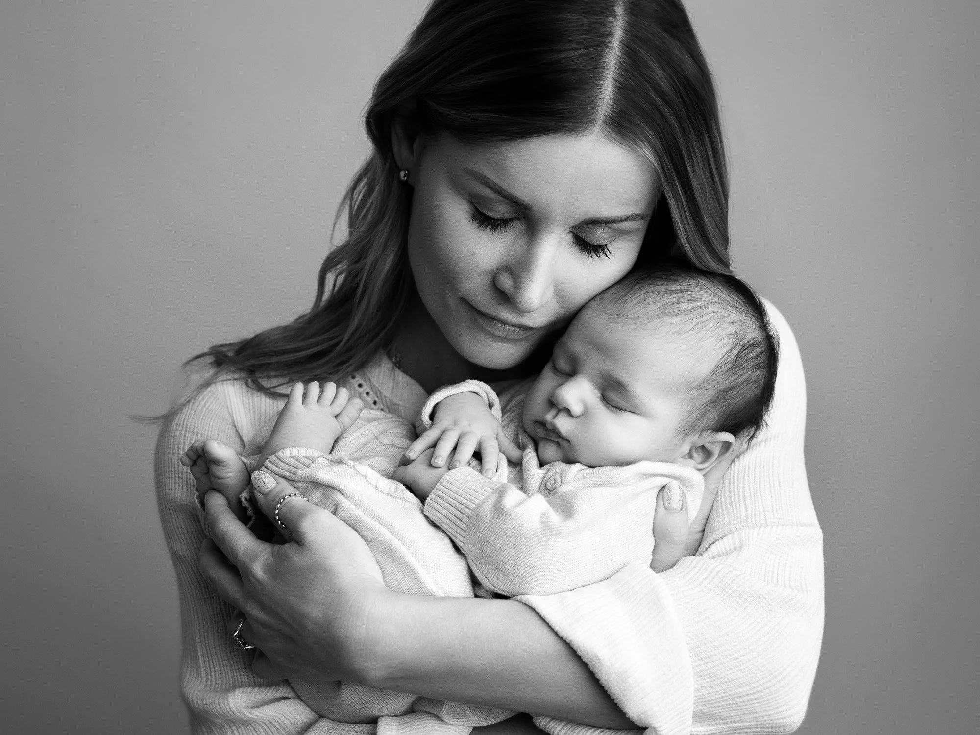 A woman holding a sleeping baby in her arms, her head resting gently on the baby's head, against a plain background.