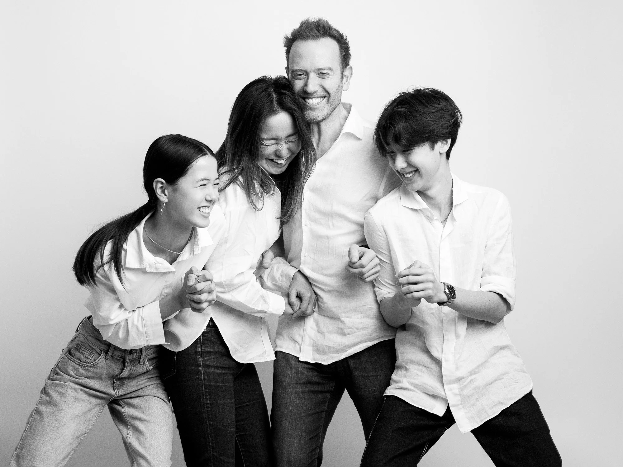 A black and white photo of a family of five laughing and holding hands, standing against a plain background.
