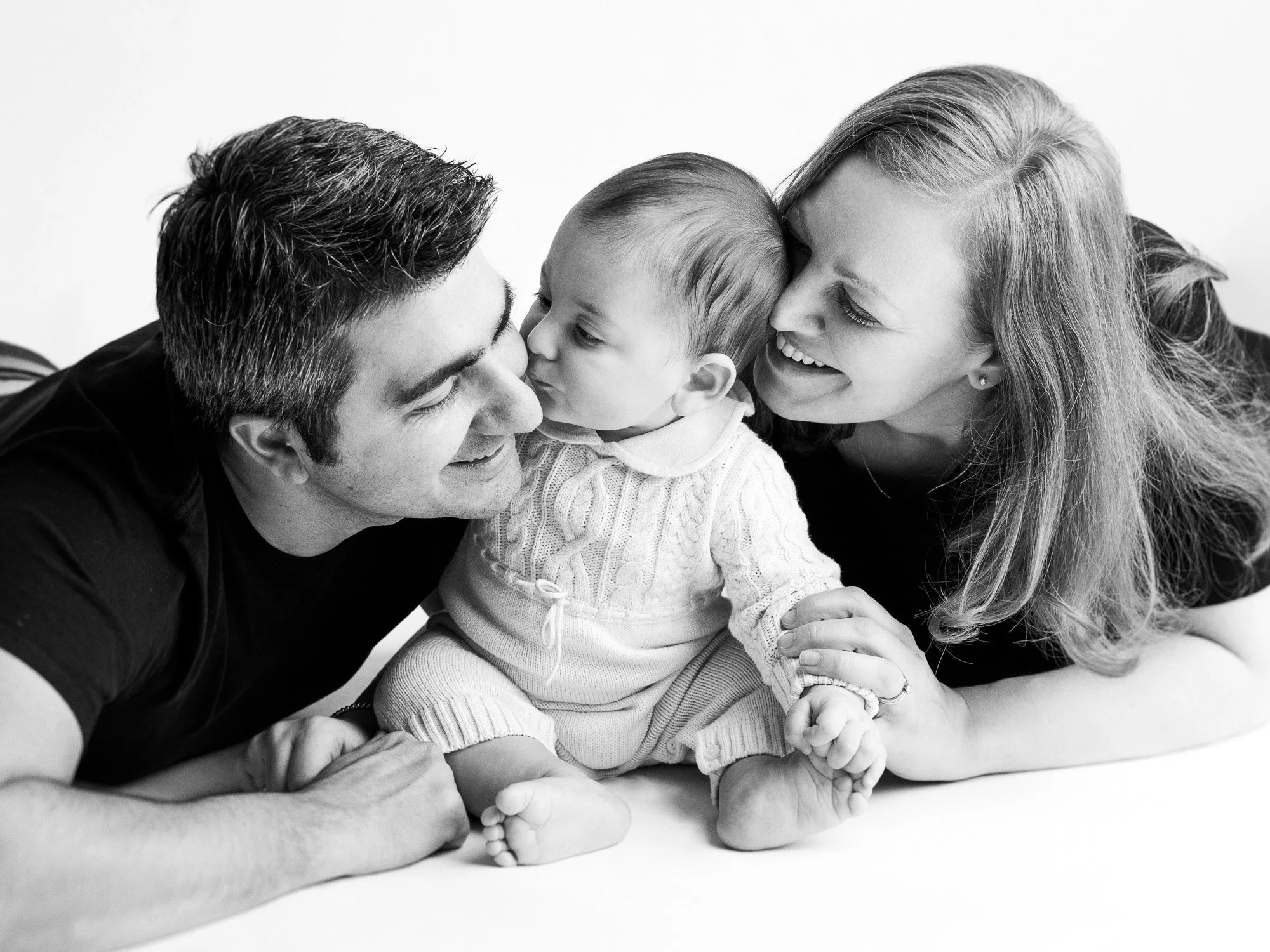 A happy family of three lying down, includes a man, a woman, and a young child. The child is in the middle, kissing the man on the nose. The woman is smiling, holding the child's hand, and close to both.