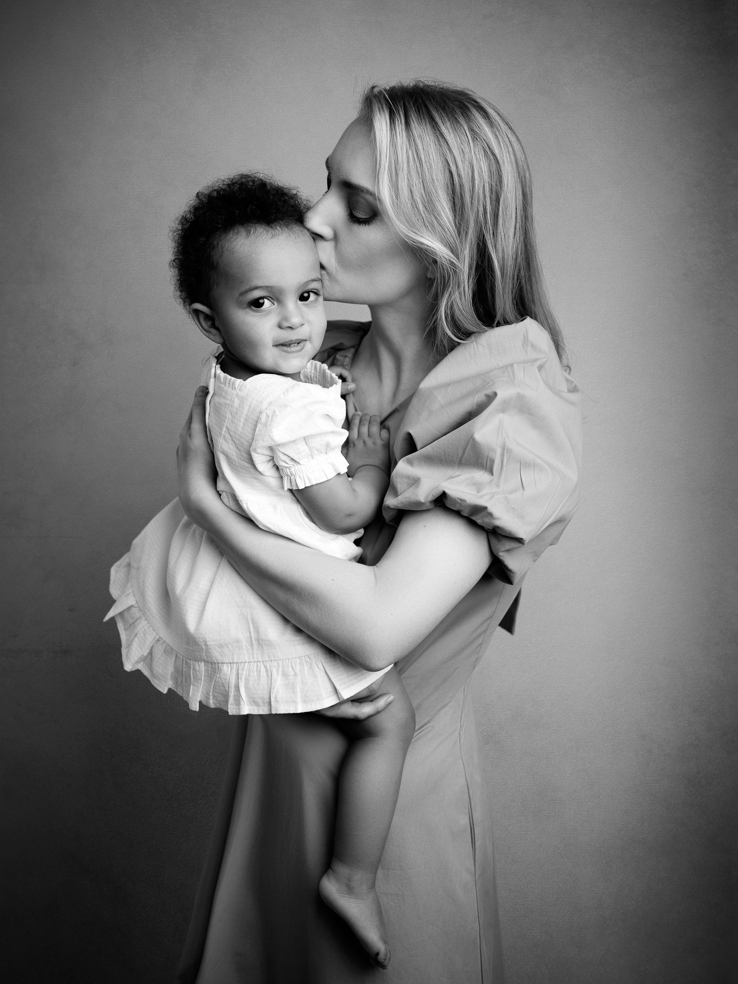 A woman with long hair holding a young girl with curly hair and wearing a white dress, with the woman kissing the girl on the forehead in a black and white photo.
