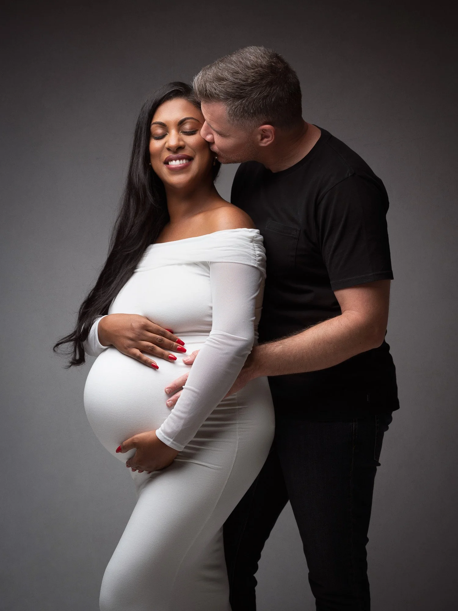 A pregnant woman in a white dress smiling with her eyes closed while a man kisses her on the cheek, against a gray background.