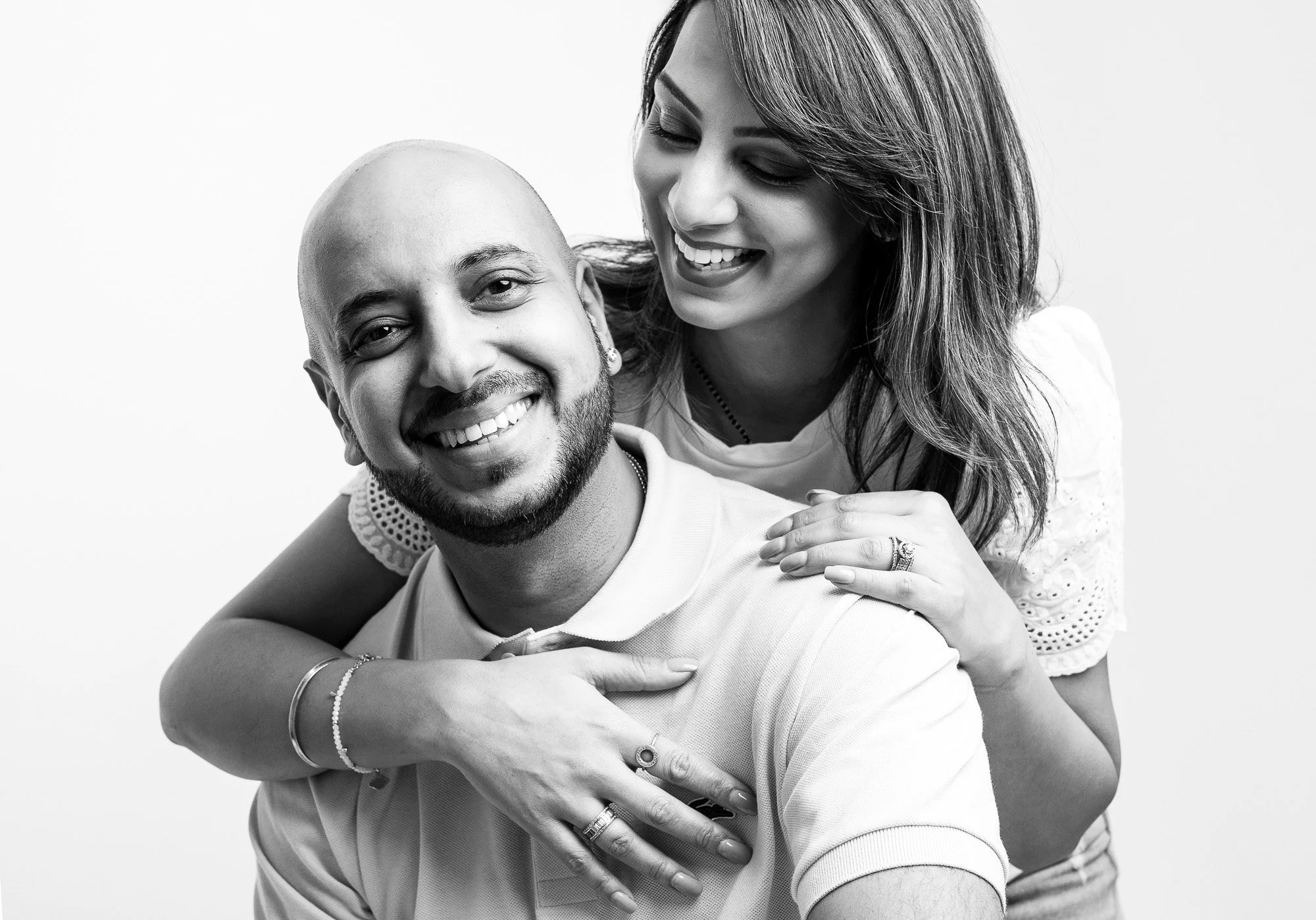 A smiling man with a bald head and a woman with long wavy hair, both in casual white shirts, sharing a joyful moment together, with the woman resting her hands on the man's shoulders.