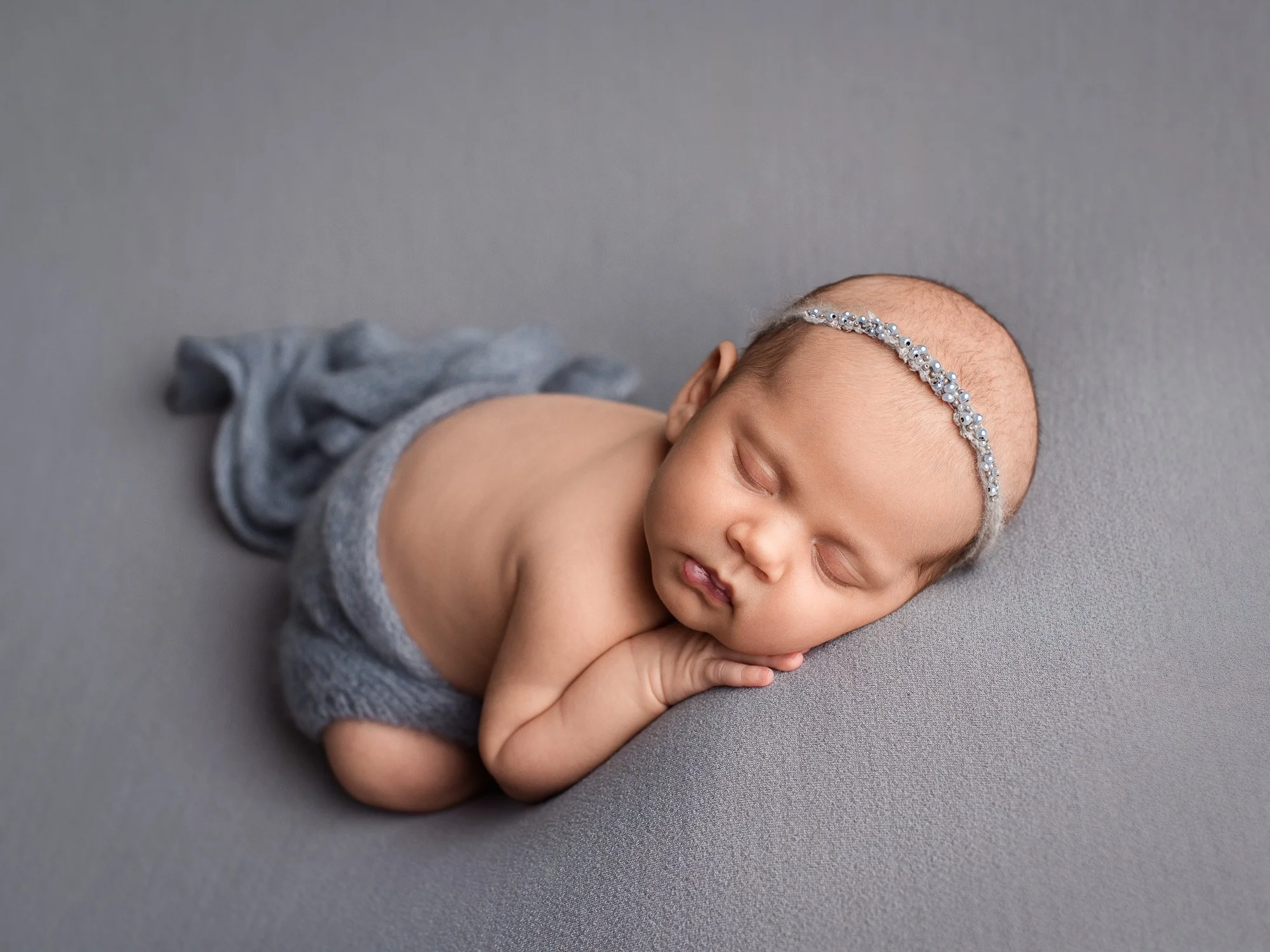A sleeping newborn baby girl with a pearl headband, lying on her side on a gray surface, wearing a gray cloth diaper.
