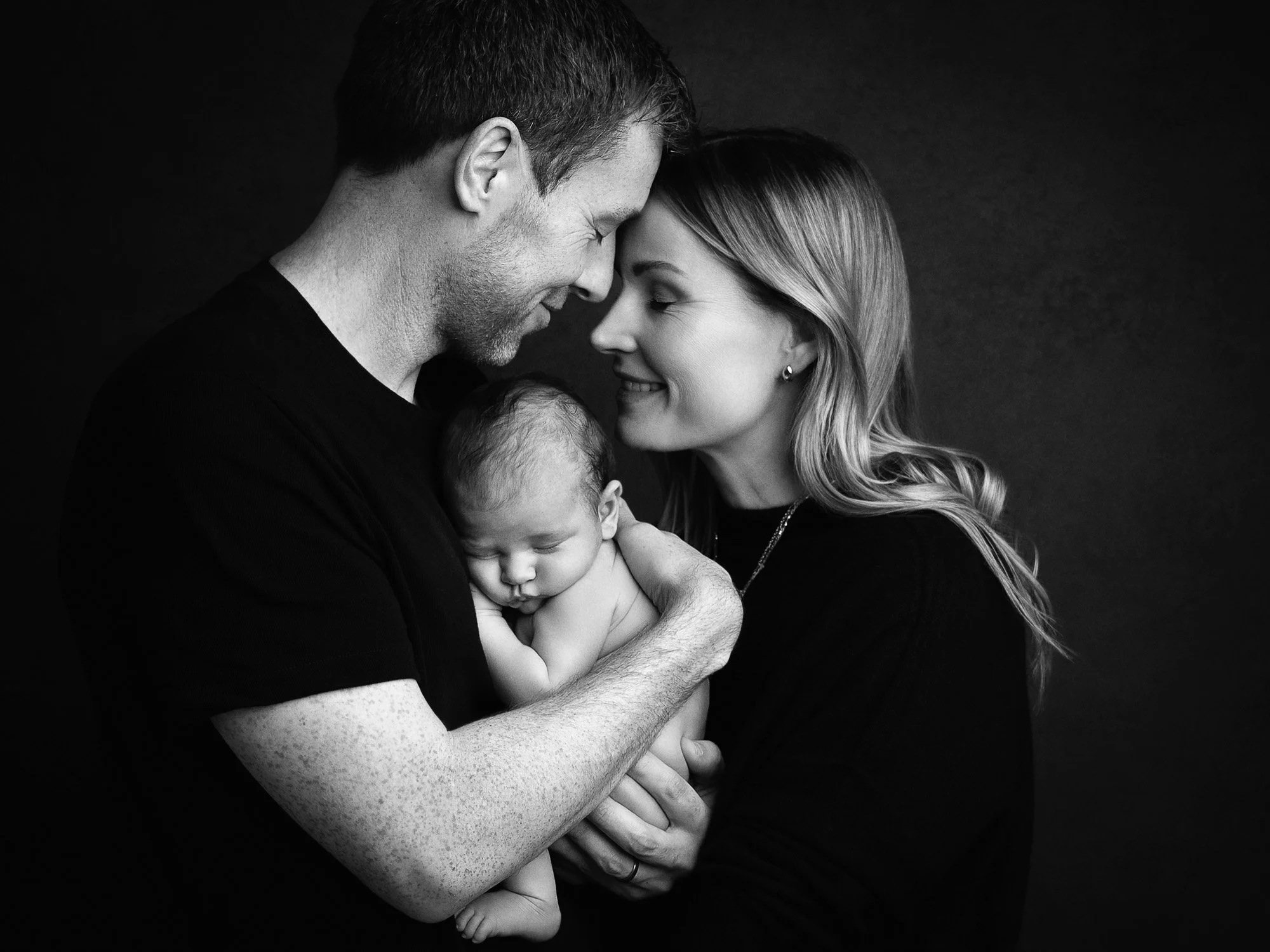 A black-and-white photo of a family of three, with a man and woman holding a newborn baby, all with their foreheads touching and smiling softly.