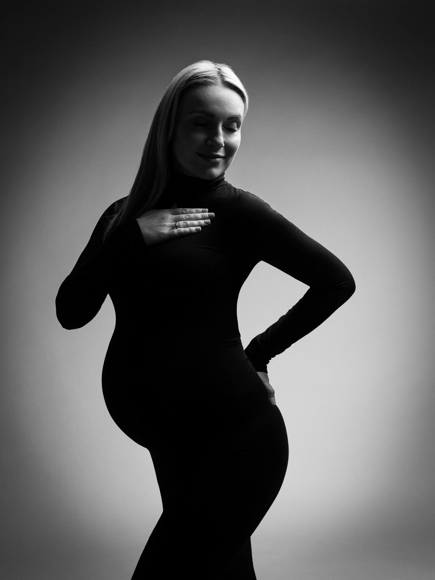 Black and white side profile of a pregnant woman with a hand on her chest, smiling gently, wearing a form-fitting dress.