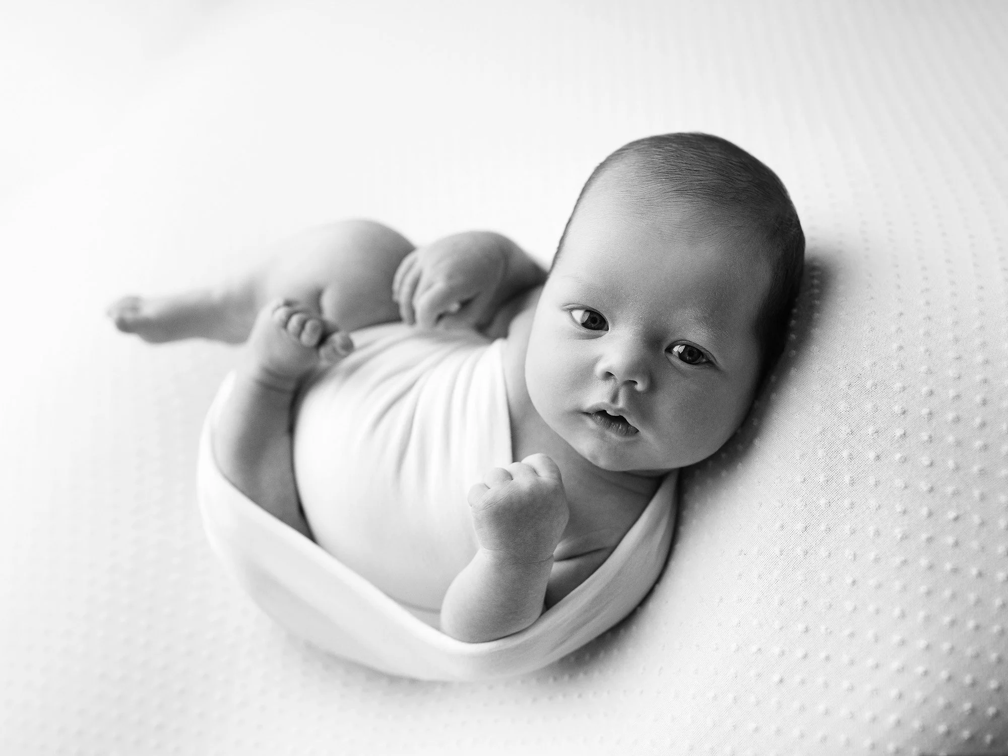 Black and white photo of a newborn baby lying on a textured surface, looking at the camera with hands near the face.