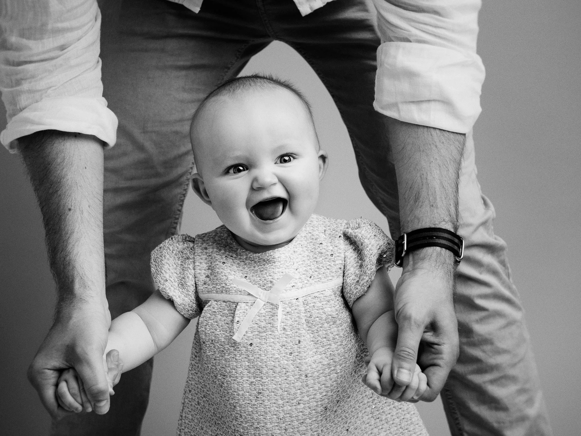 A smiling baby girl with short hair, wearing a textured dress with a small bow, holding hands with an adult, who is partially visible, against a plain background.