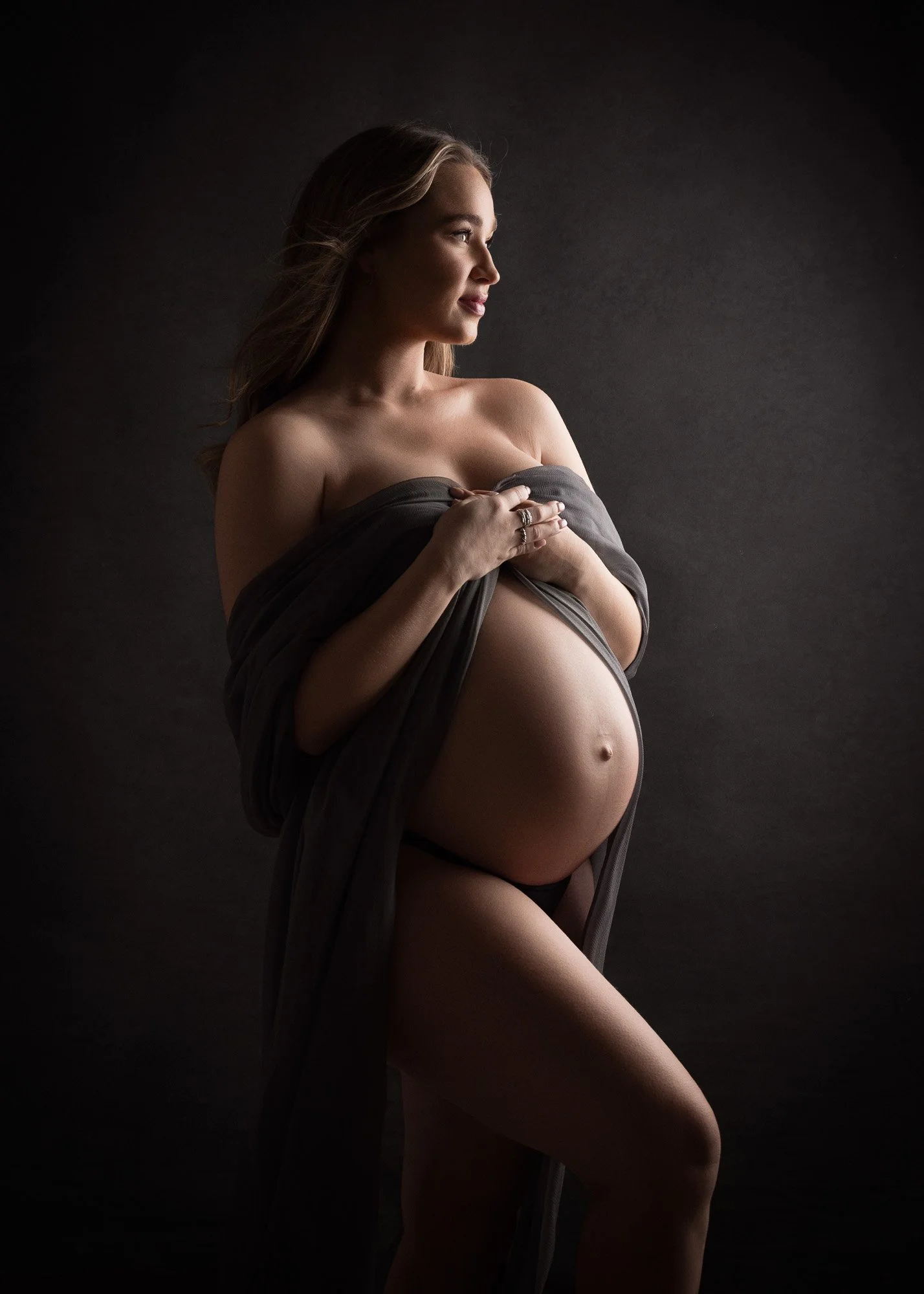 A pregnant woman with long hair, wrapped in dark fabric, standing against a dark background. She is looking to the side and holding her belly.