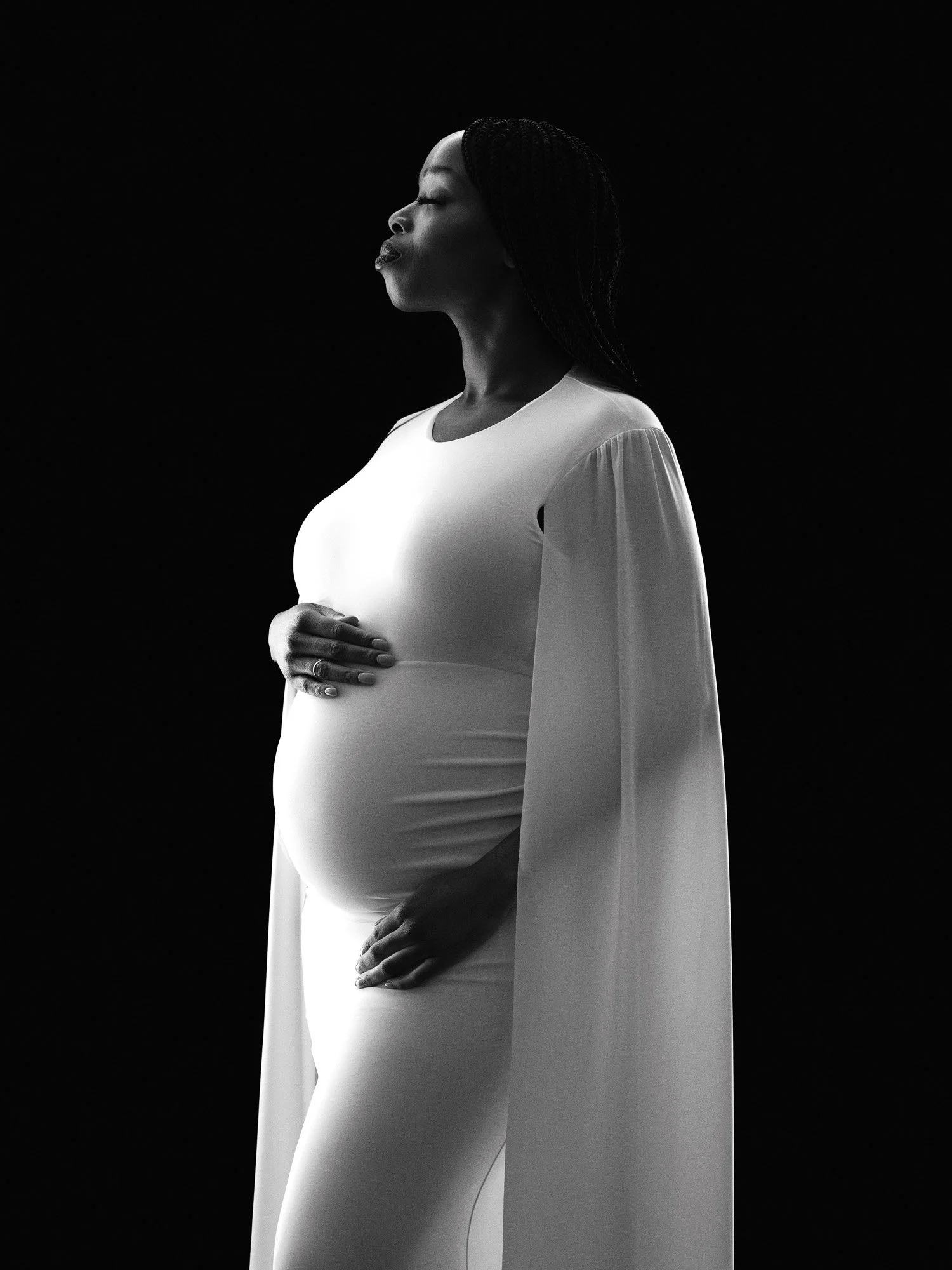 A pregnant woman with braided hair wearing a white dress with long, flowing sleeves, standing in profile against a black background.