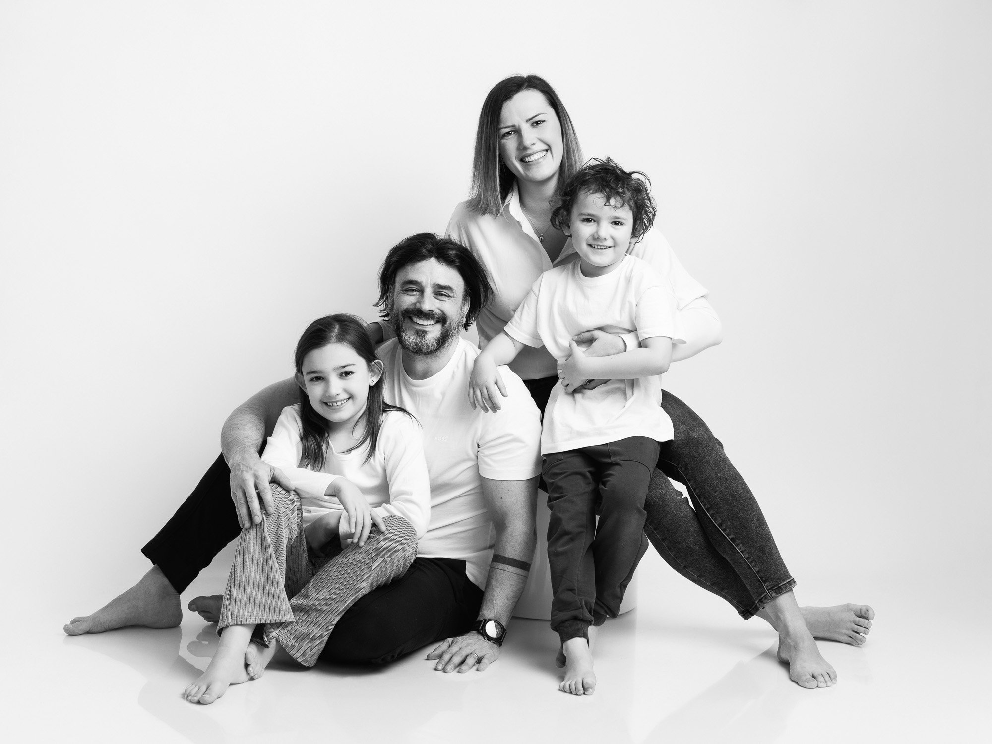 Black and white photo of a happy family of five posing together against a plain background. The father is sitting on the floor, smiling, with a girl sitting on his lap. The mother is standing in the back with her arms around a boy sitting on her lap,