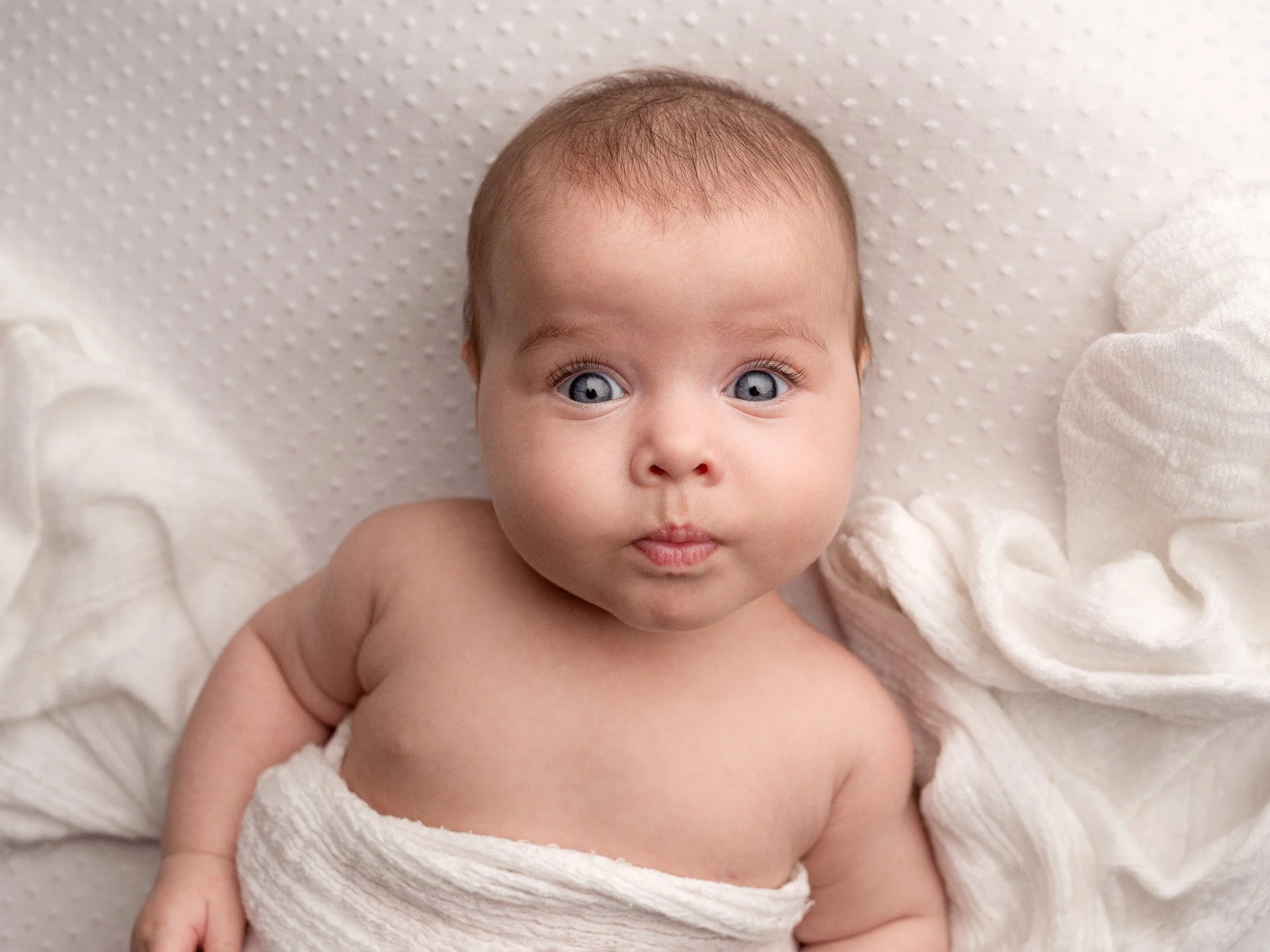 Close-up of a baby with blue eyes, light brown hair, and fair skin, lying on a white dotted blanket, wrapped in a soft white cloth, looking directly at the camera.